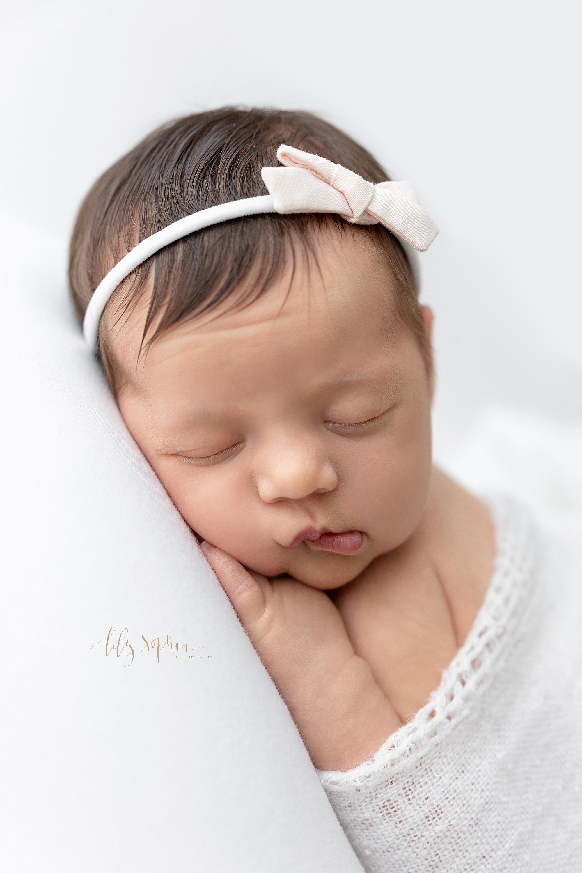  Newborn portrait of a newborn baby girl as she wears a stretchy headband with a bow as she lies on her stomach with her left hand under her cheek taken in a natural light studio near Virginia Highlands in Atlanta, Georgia. 