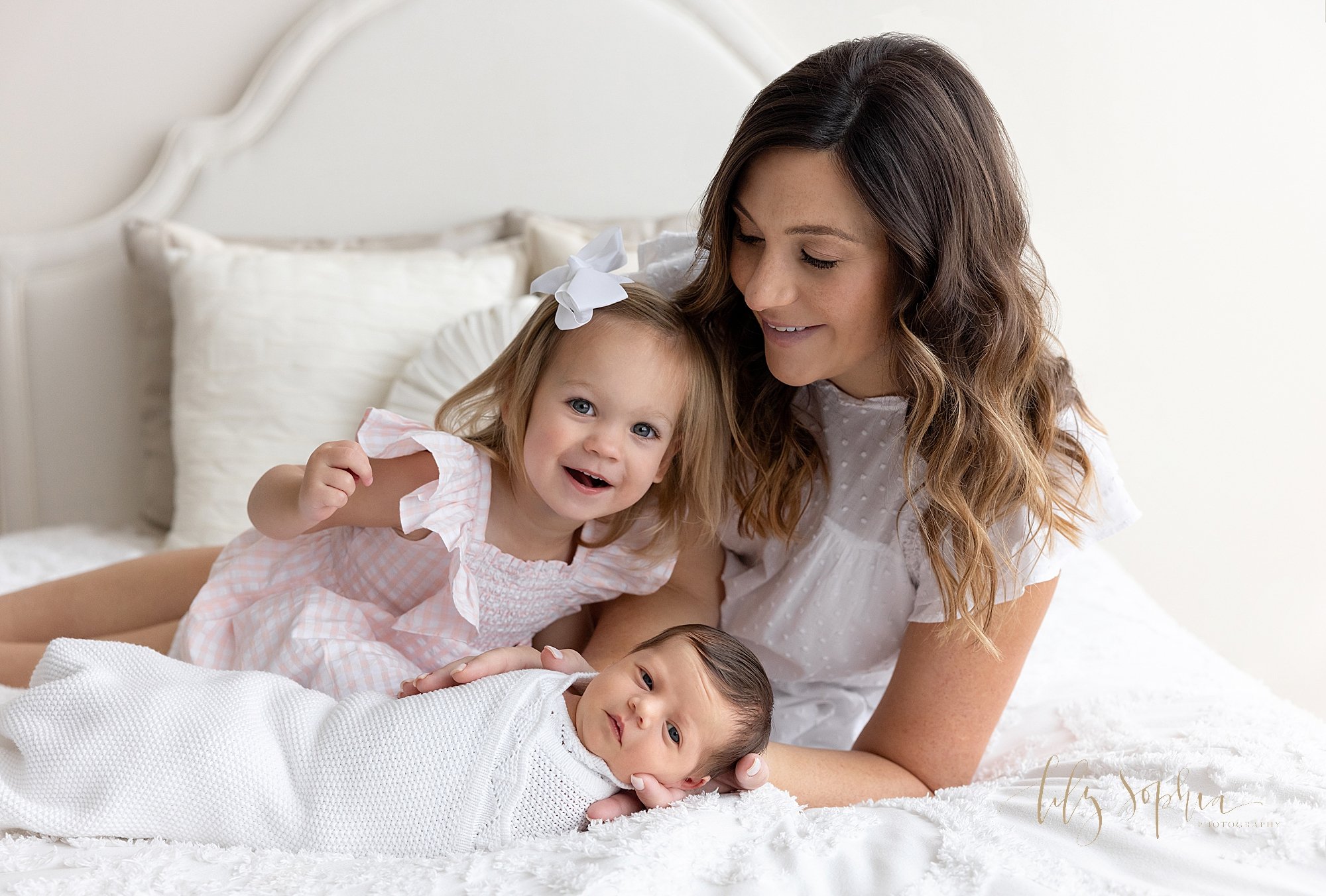  Newborn family photo of a mother lying to support herself with her left arm as she cradles the head of her newborn daughter in her right hand and as her toddler daughter sits between them taken near Buckhead in Atlanta in a photography studio that u