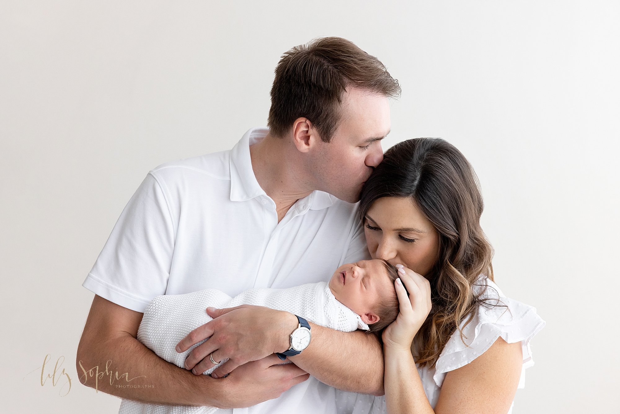  The love of a family is captured in this newborn photo of a father cradling his newborn daughter in his arms as mom stands to his right side kissing the head of her daughter and placing her left hand on her daughter’s head as her husband kisses her 