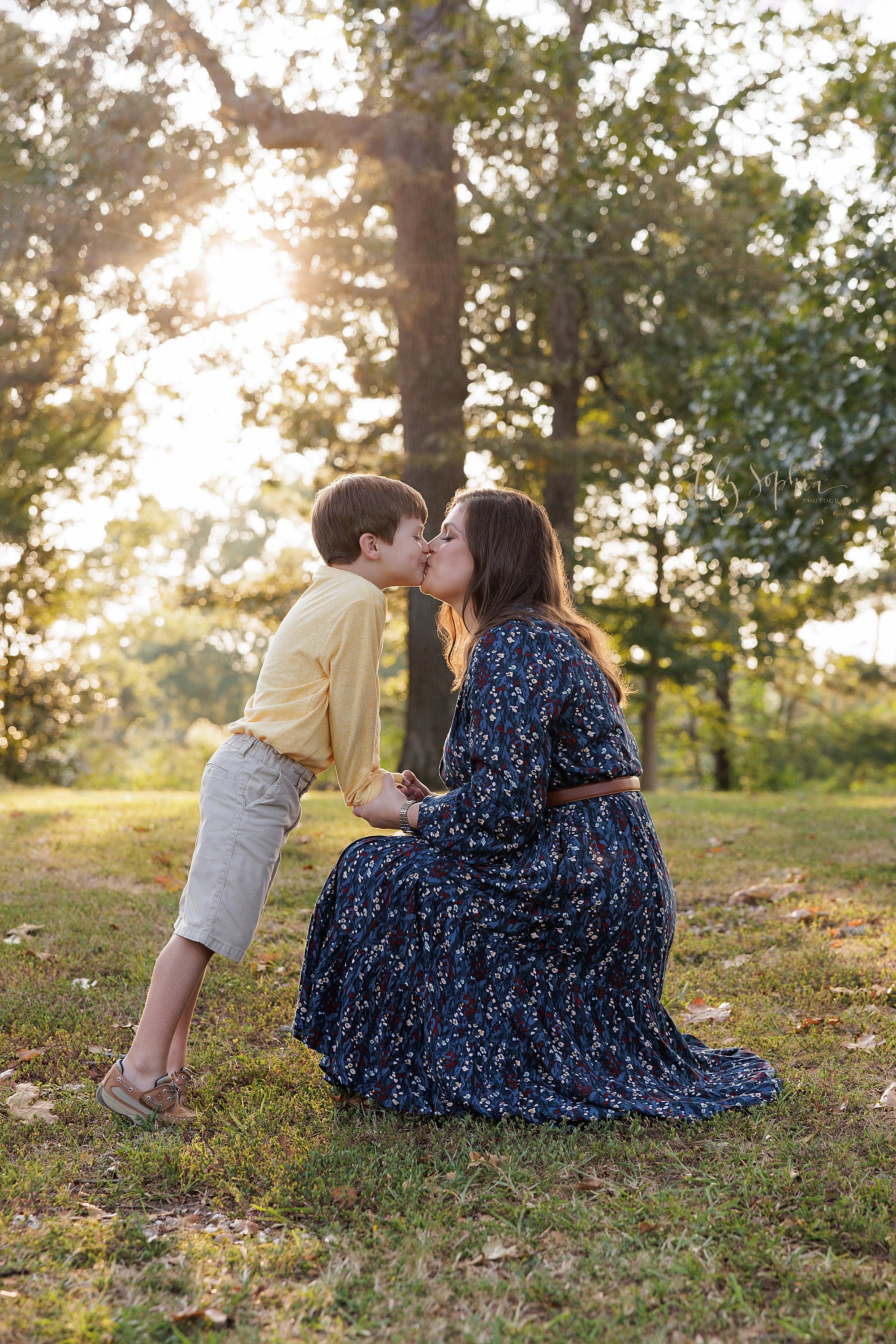  Mother and son portrait taken at sunset in a park near Atlanta, Georgia with mom squatting so she can kiss her son who faces her and holds her hands. 