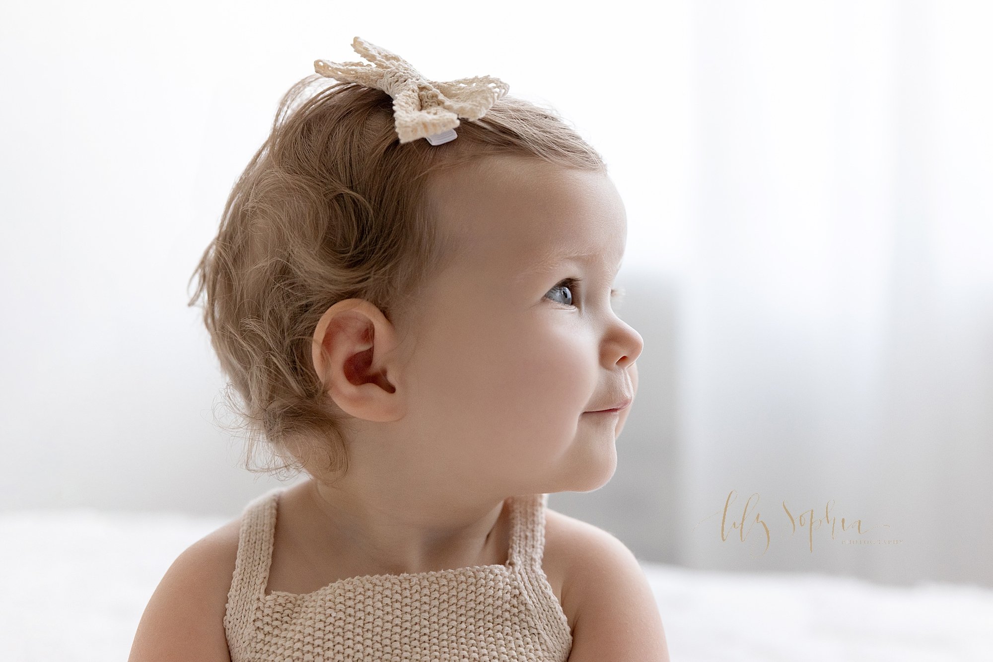  First birthday profile portrait of a one year old baby girl as she turns to look over her right shoulder as she sits in front of a window streaming natural light in a photography studio near Oakhurst in Atlanta. 