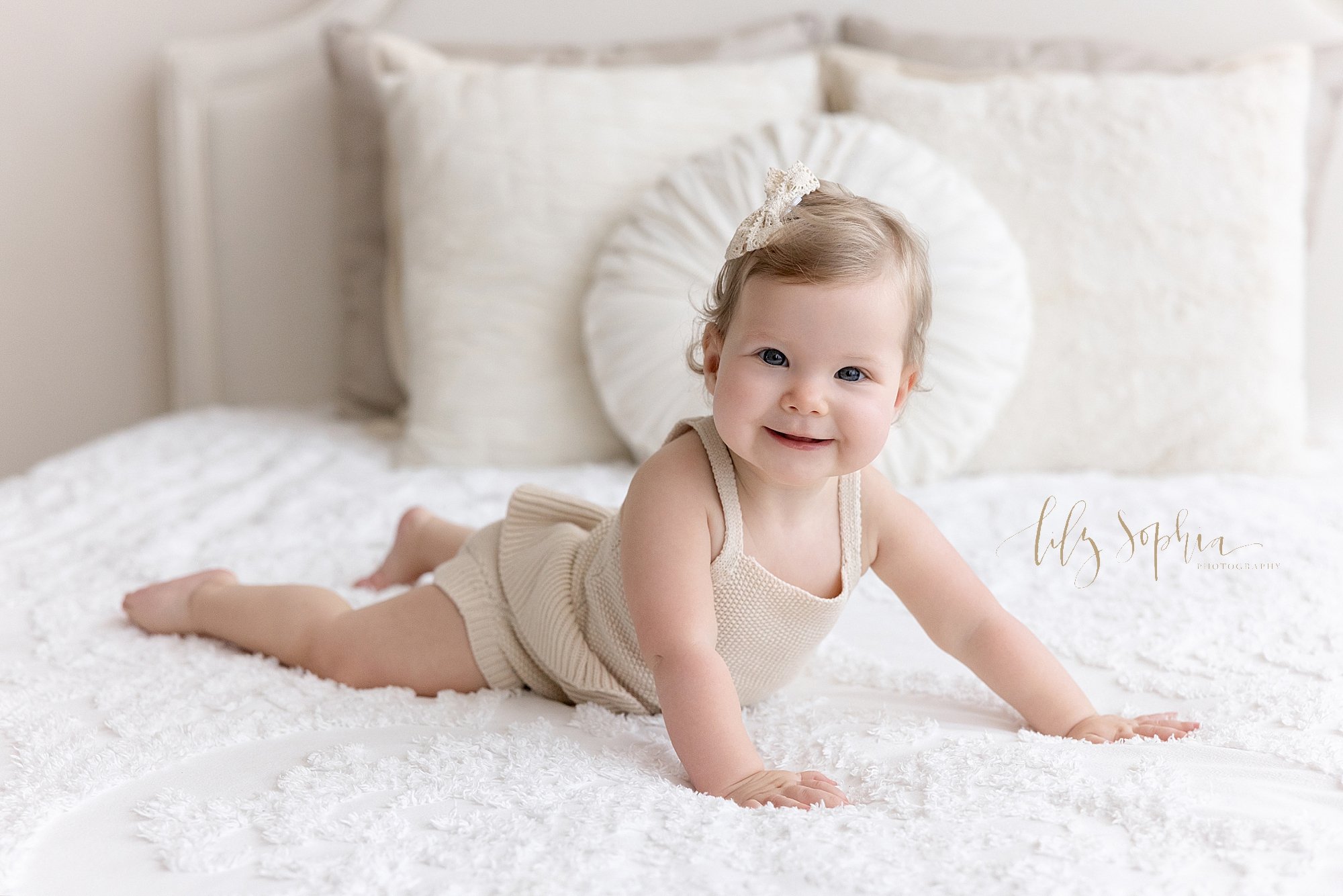  Portrait of a smiling one year old baby girl as she lies on her stomach on a bed and pushes her self up with her arms taken for her first birthday in a natural light studio near Ansley Park in Atlanta. 