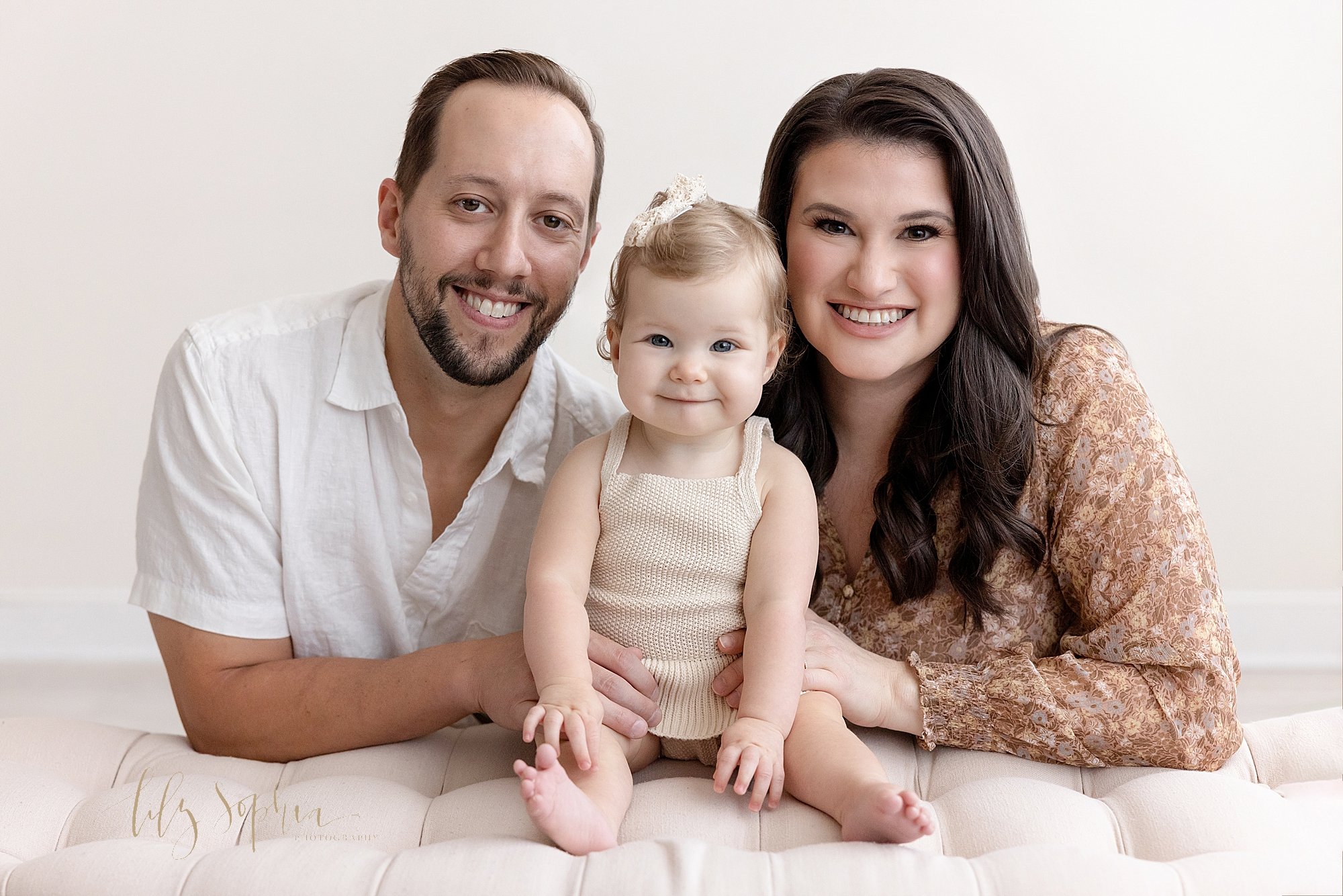  Family portrait of a mother and father kneeling behind a tufted bench as their one year old daughter sits between them taken using natural light in a studio near Kirkwood in Atlanta, Georgia. 