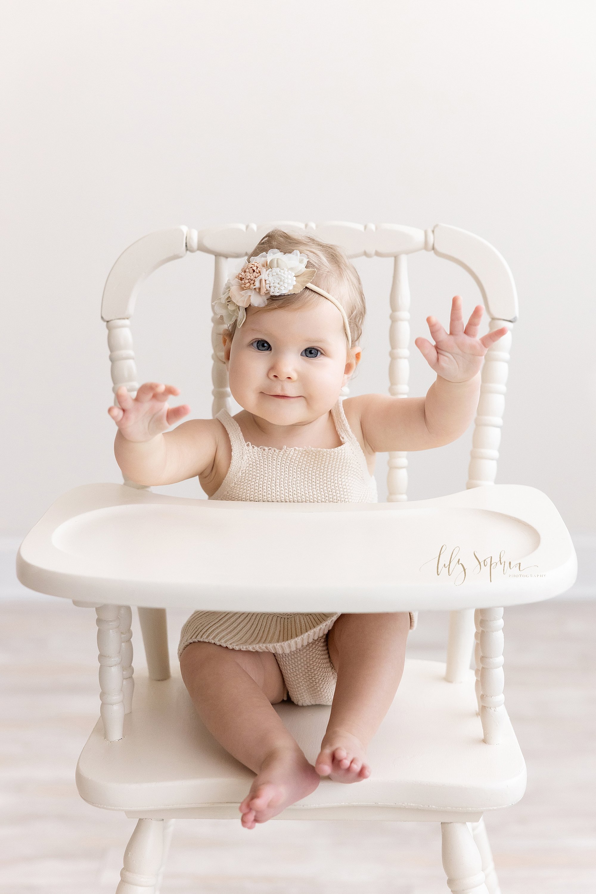  First birthday photo session of an excited one year old baby girl as she sits in an antique high chair and raises her arms taken near Vinings in Atlanta in a natural light photography studio. 