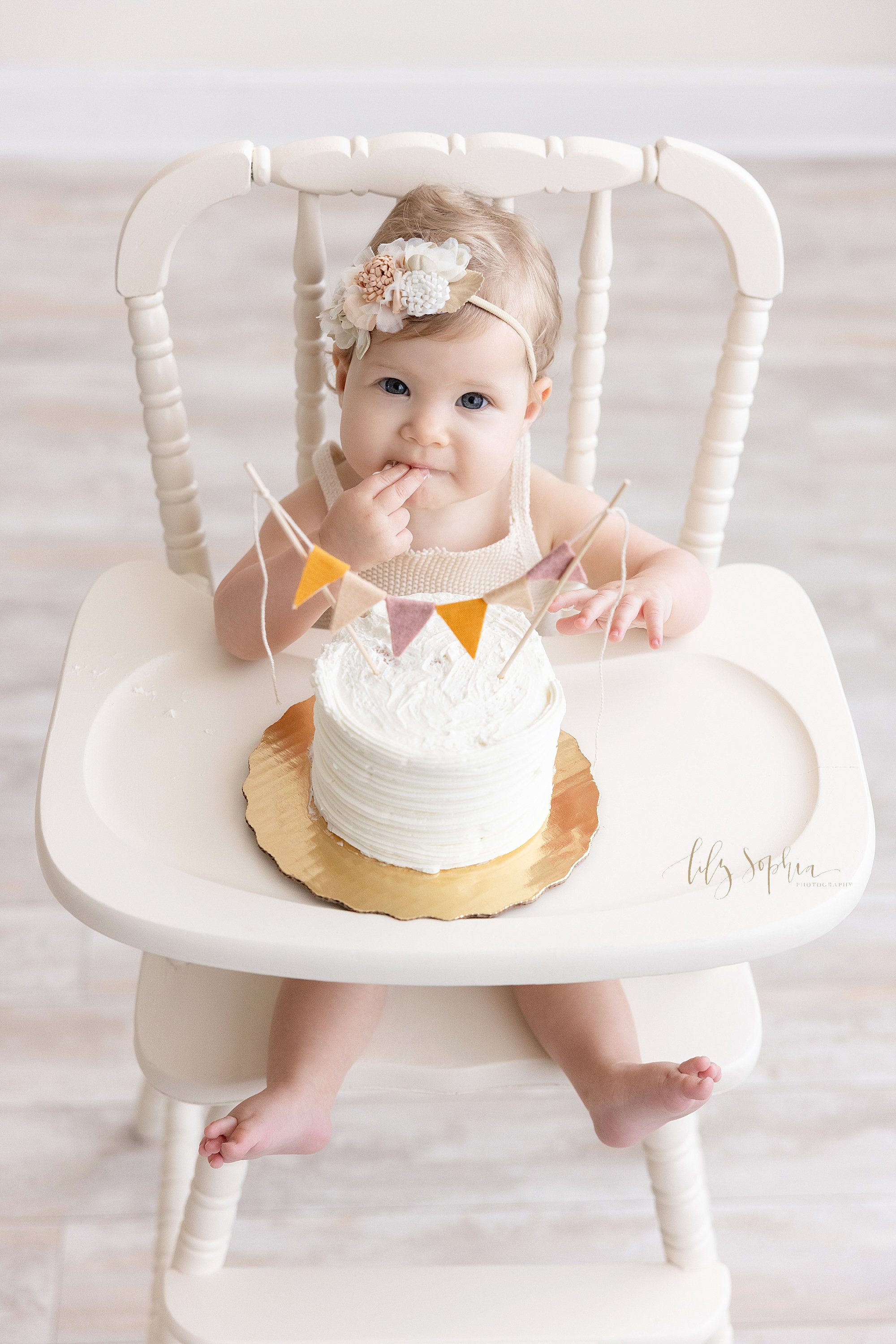  First birthday smash cake portrait of a one year old baby girl as she daintily tastes the icing of her smash cake with the fingers of her right hand taken in natural light in a studio near Midtown in Atlanta. 