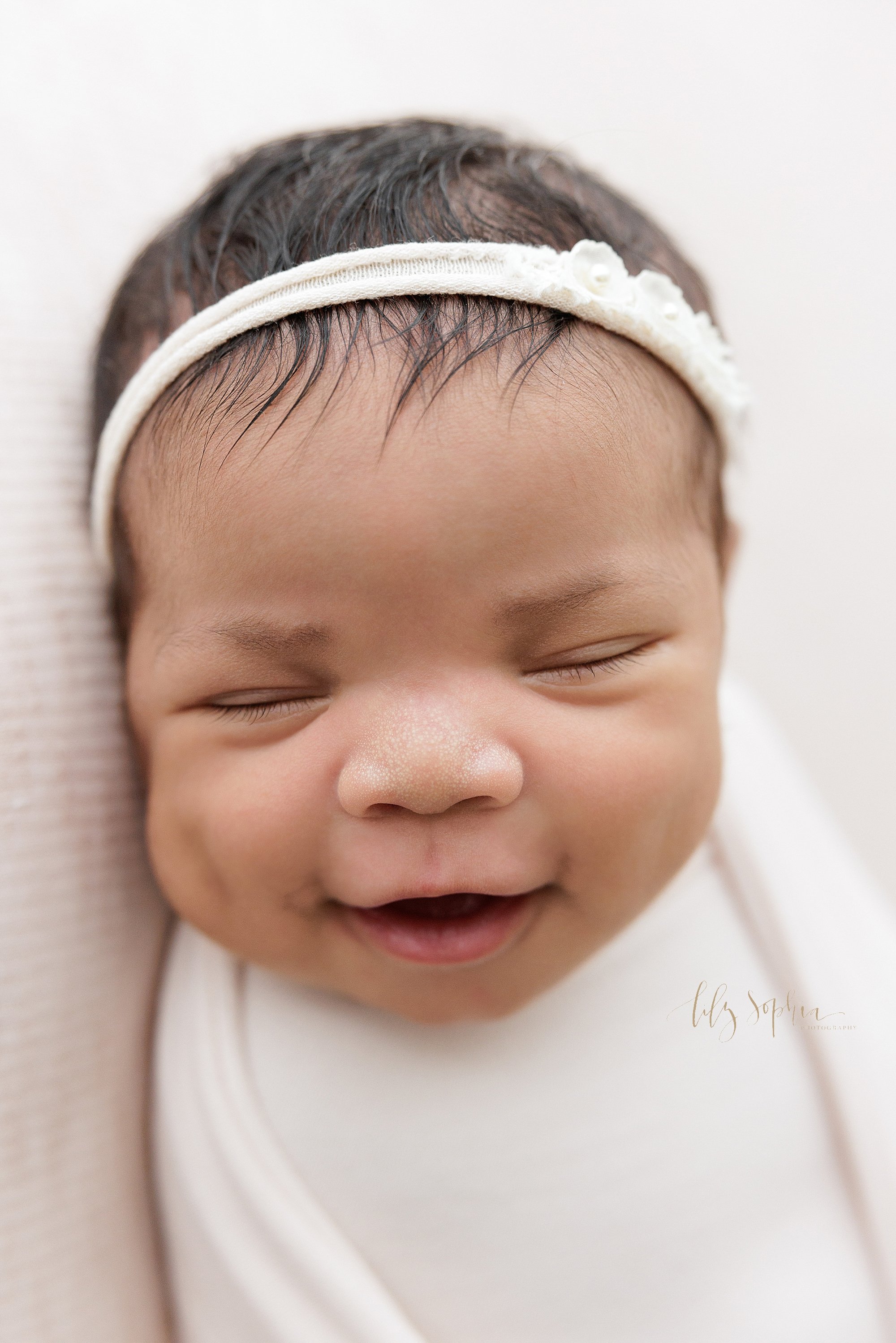  Newborn close-up portrait of a newborn African-American baby girl wearing a stretchy headband in her hair as she lies swaddled to her neck and smiles in her sleep taken near Virginia Highlands in Atlanta, Georgia in a natural light studio. 