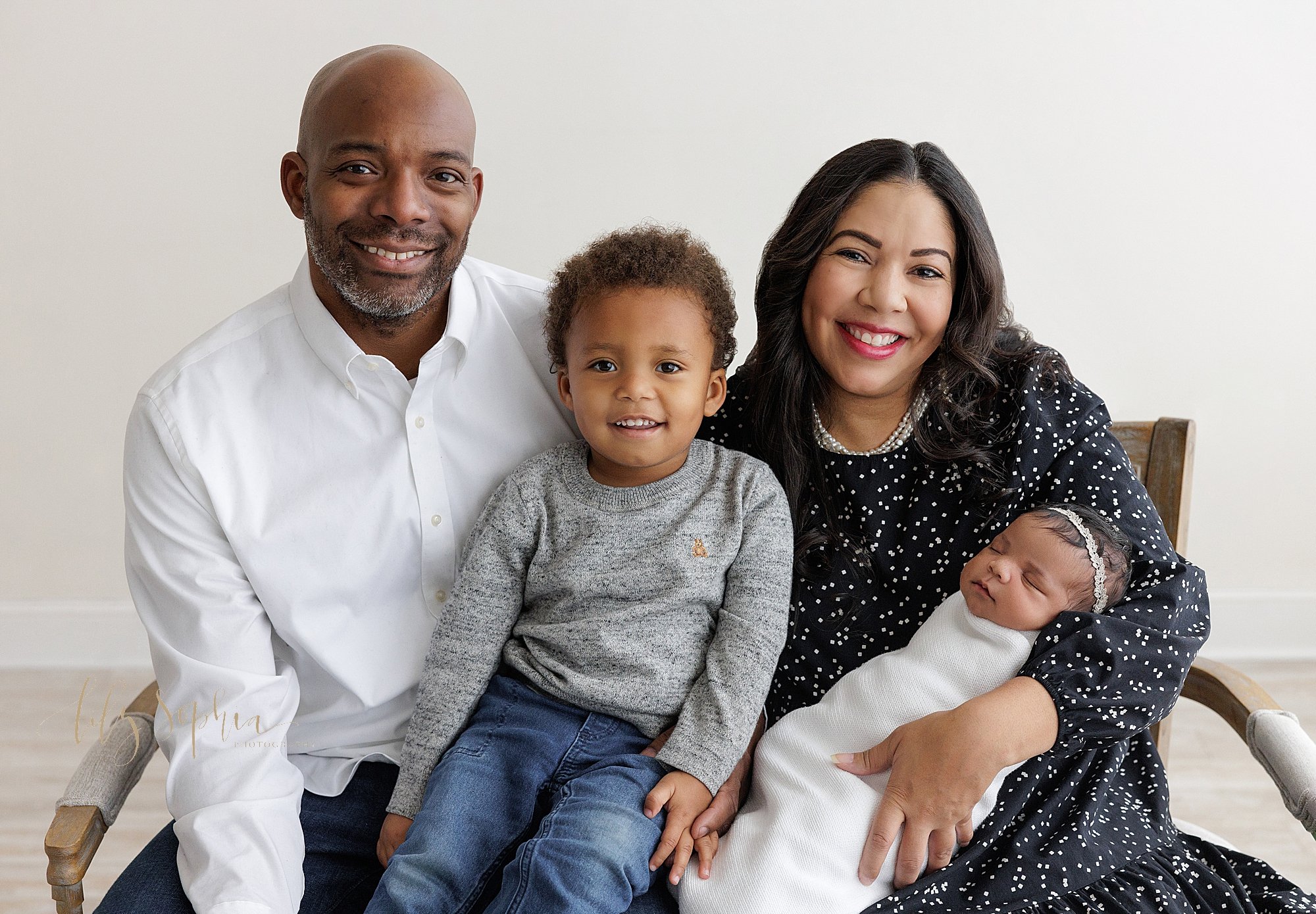  Family newborn photo of a mother sitting and holding her newborn daughter in her left arm as her husband sits next to her holding their young son on his lap taken near Roswell in Atlanta in a natural light studio. 