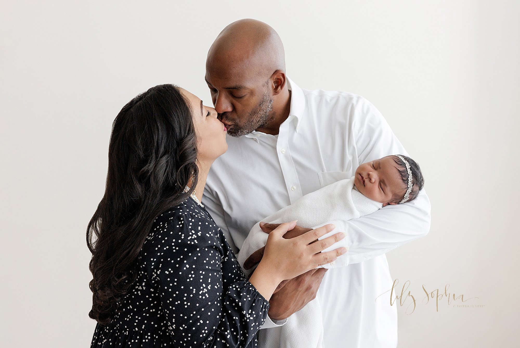  The love of a husband and wife is captured in this newborn photo of a father cradling his newborn daughter in his left arm as his wife stands next to him and the two of them kiss taken near Smyrna in Atlanta in a studio using natural light. 