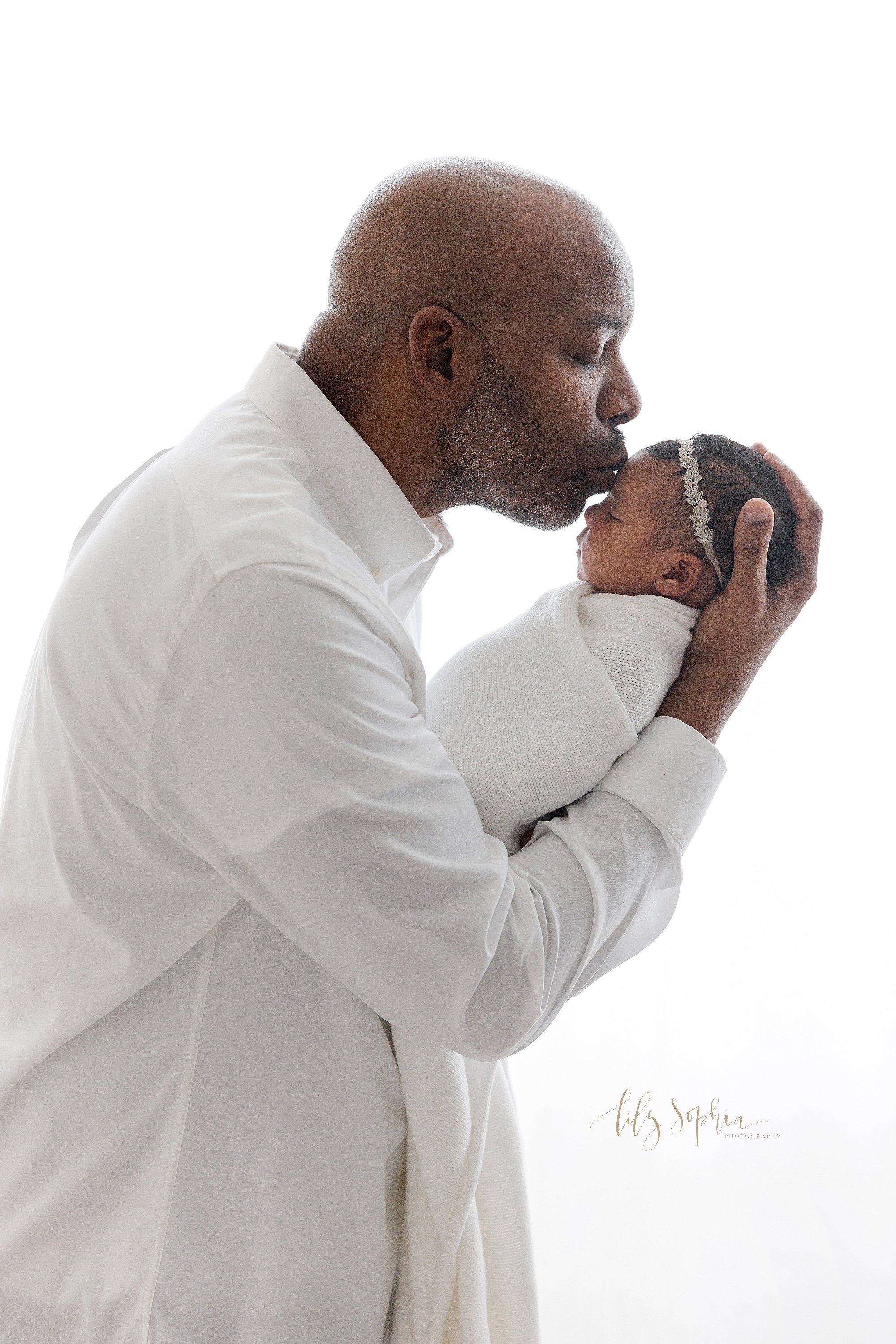  Portrait of an African-American father as he holds his newborn baby daughter in his hands and kisses her forehead taken in front of a window streaming natural light in a photography studio near Morningside in Atlanta, Georgia. 