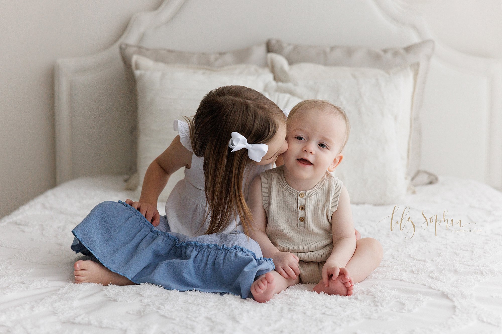  Precious sibling photo of a one year old baby boy as he sits next to his older sister on a bed and she kisses him on his cheek taken in a photography studio that uses natural light near Alpharetta in Atlanta. 