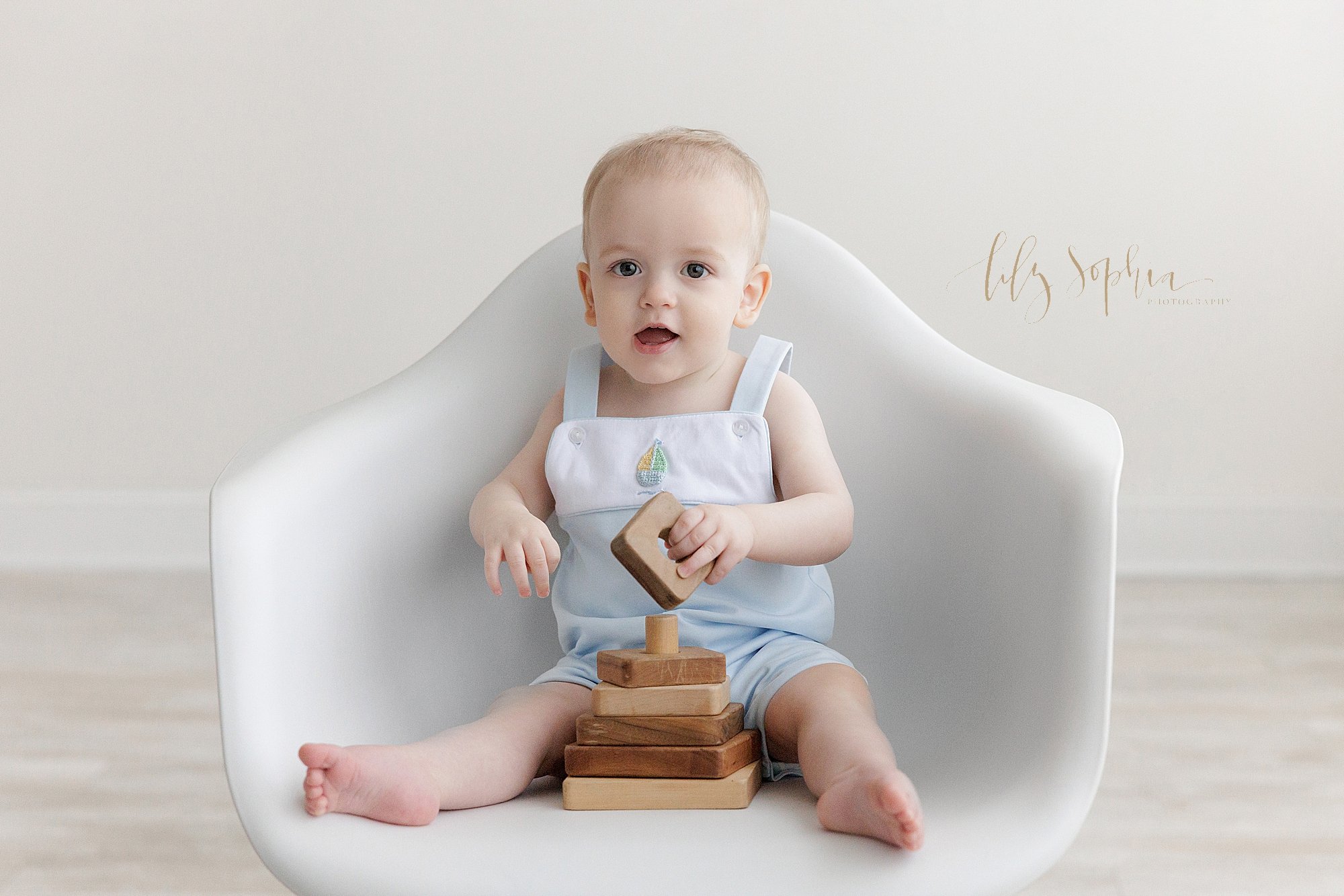  A one year old baby boy sits in a molded white chair playing with a wooden stacking toy for his first birthday portrait taken in a studio using natural light near Brookhaven in Atlanta, Georgia. 