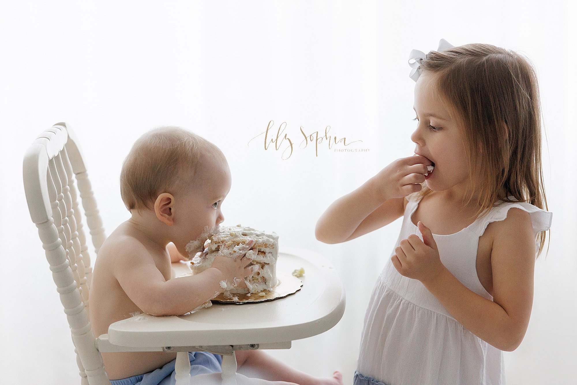  Smash cake sibling photo of a one year old baby boy sitting in an antique high chair holding on to his cake and biting into it as his older sister faces him and stuffs icing into her mouth taken in front of a window streaming natural light into a ph