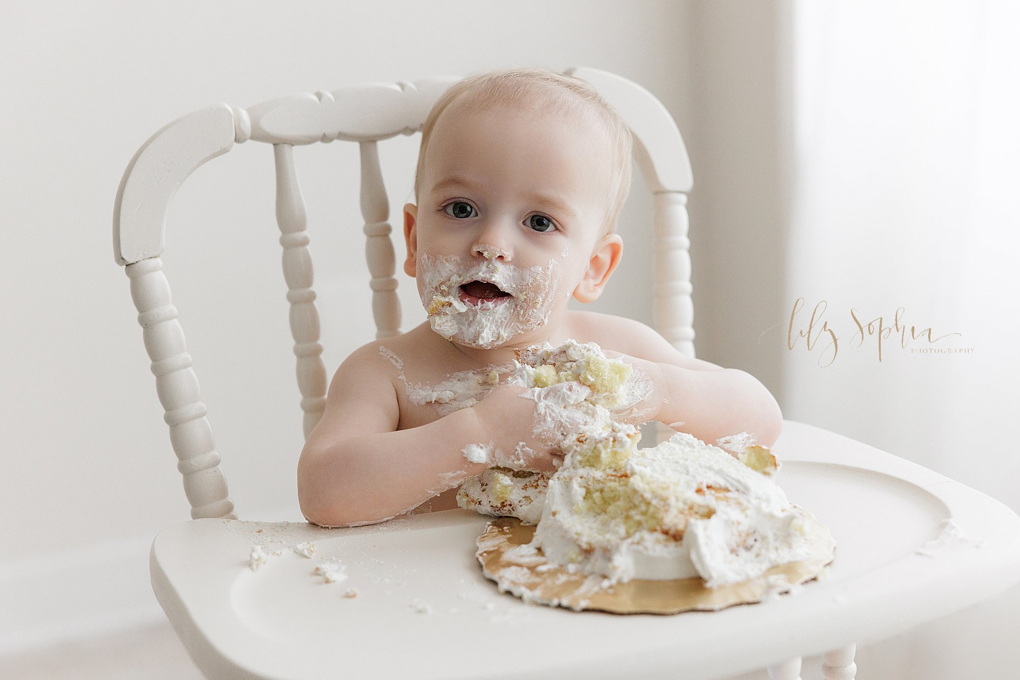  First birthday smash cake photo session with a happy one year old baby boy as he sits next to a window streaming natural light in an antique high chair with his face covered in icing, his hands clutching cake and icing, and his smash cake destroyed 