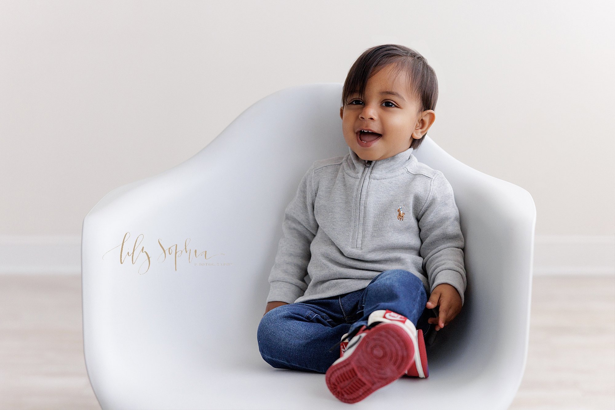  A proud Indian boy smiles as he sits in a white molded chair on his first birthday during a photo session in a photography studio that uses natural light near Brookhaven in Atlanta. 