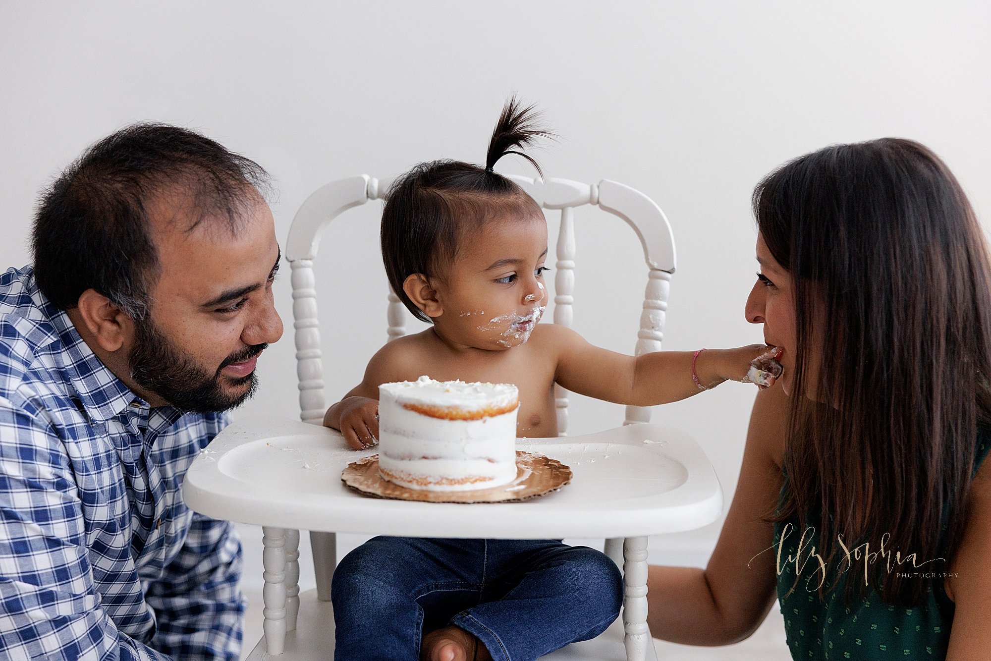  Surrounded by his parents as they squat next to him a one year old little boy sits in an antique high chair and feeds his mom icing from his smash cake as he celebrates his first birthday having a photo session near Smyrna in Atlanta in a natural li