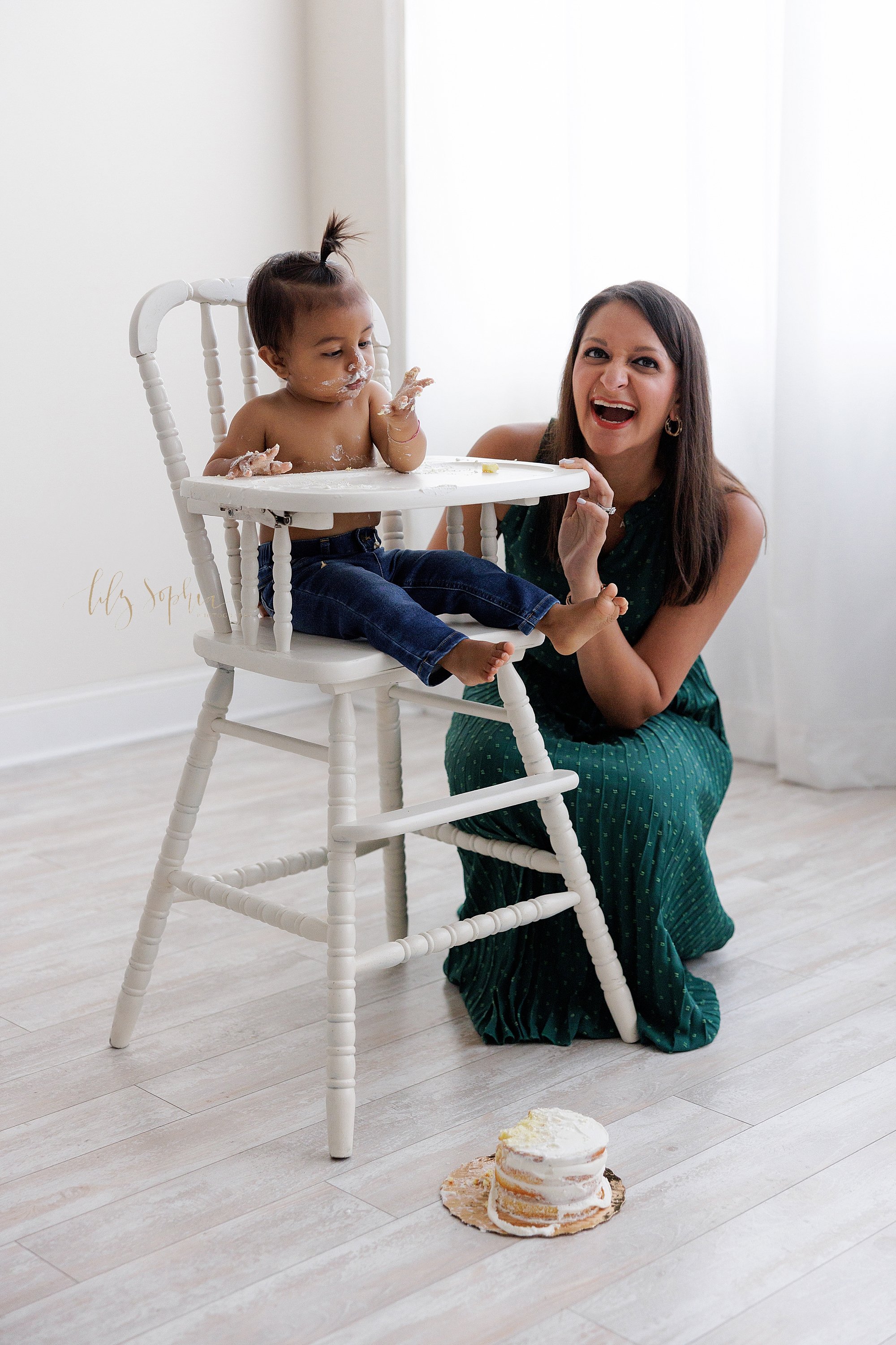 A one-year old little boy sits in an antique highchair covered in icing that is left from his smash cake for his first birthday as his mom squats next to him and laughs taken in front of a window streaming natural light in a photography studio near 