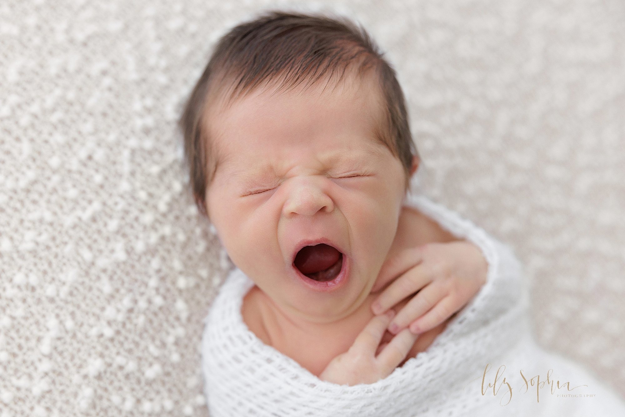  Newborn picture of a newborn baby girl wrapped in a soft white knitted blanket as she yawns to wake up taken in Ponce City Market in Atlanta, Georgia in the Lily Sophia Photography Studio. 
