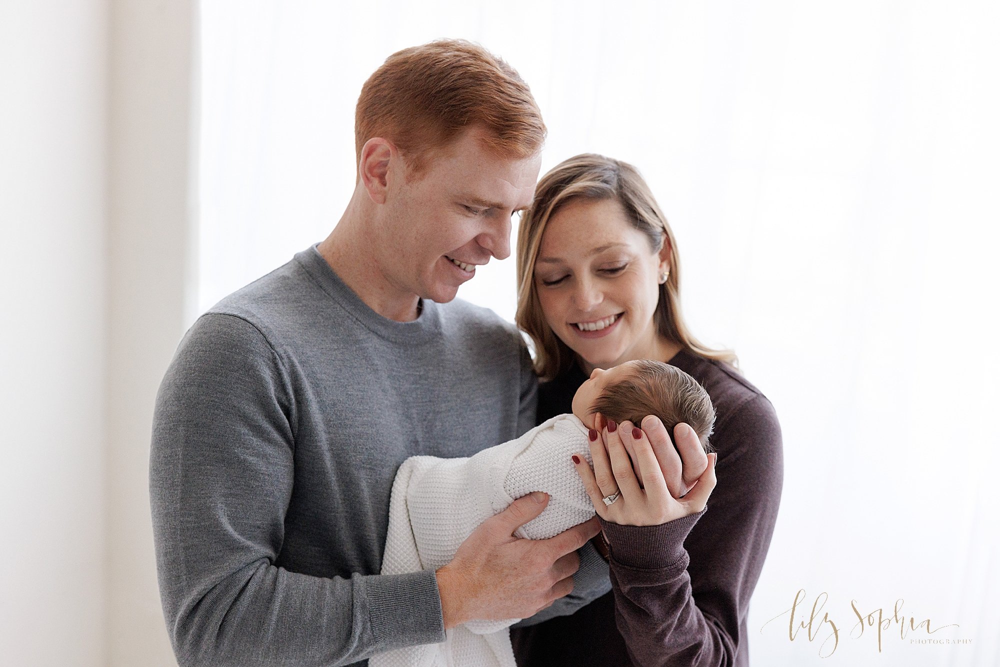  Picture of a father holding his newborn baby girl in his arms in front of his chest as his wife stands to his right side and places her left hand on his hand to hold their daughter’s head and the couple admire their newborn daughter while standing i
