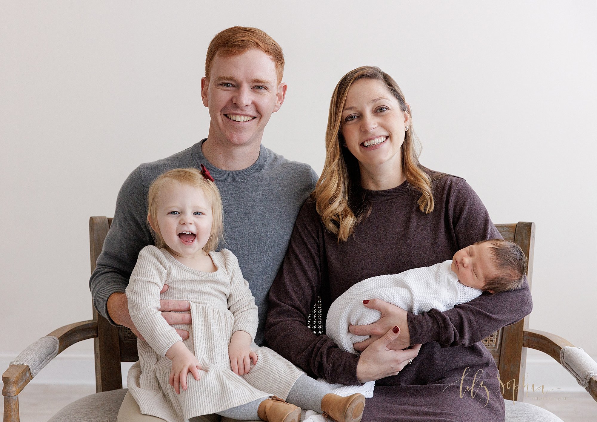  Family newborn photo of a mom cradling her sleeping newborn baby girl in her left arm as her husband sits next to her with their toddler daughter on his lap taken near Old Fourth Ward in Atlanta in a photography studio that uses natural light. 