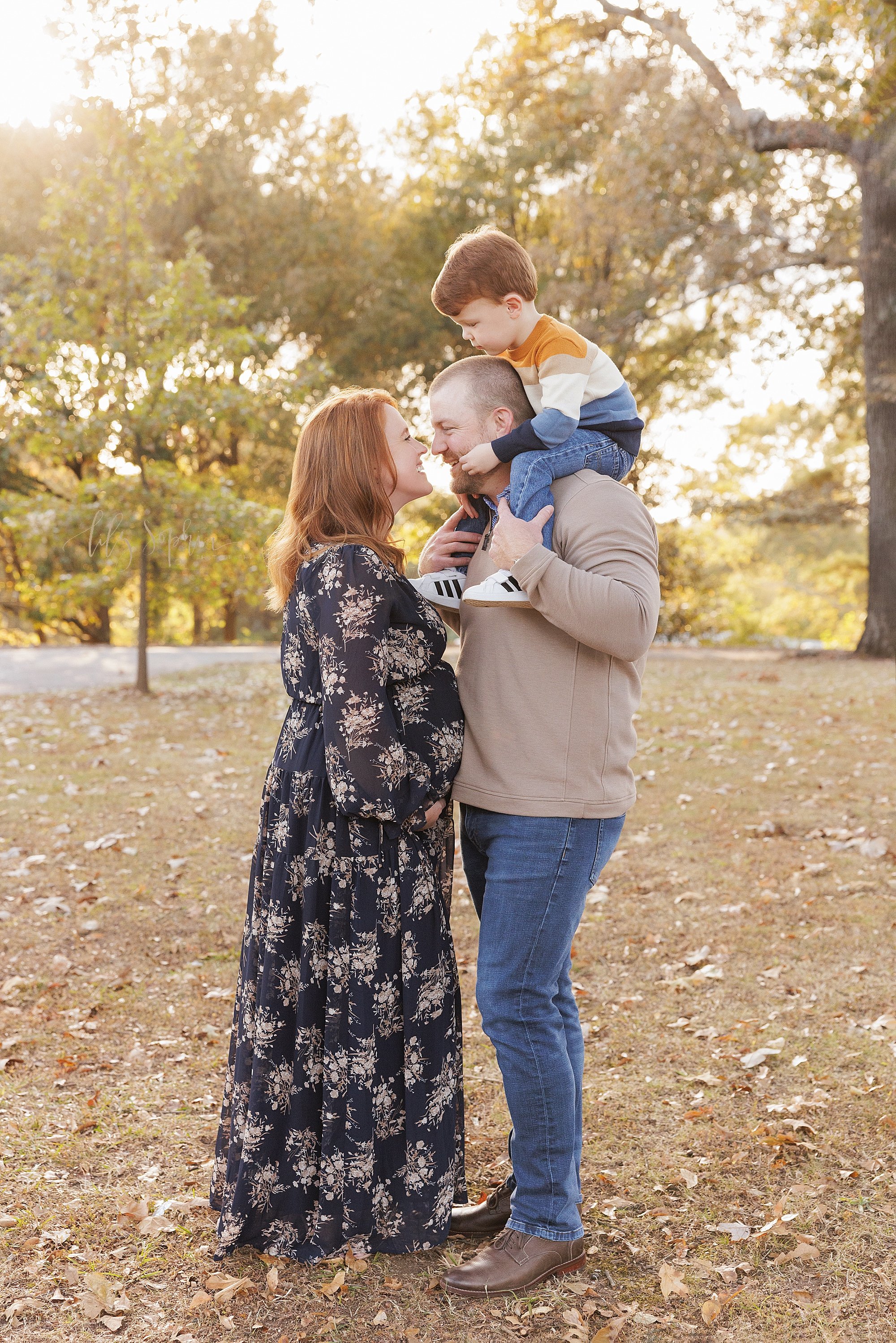  Maternity photo session in a park near Atlanta, Georgia of a pregnant mother holding the base of her belly as she faces her husband who is holding their toddler son on his shoulders during sunset in autumn as they get ready to kiss one another. 
