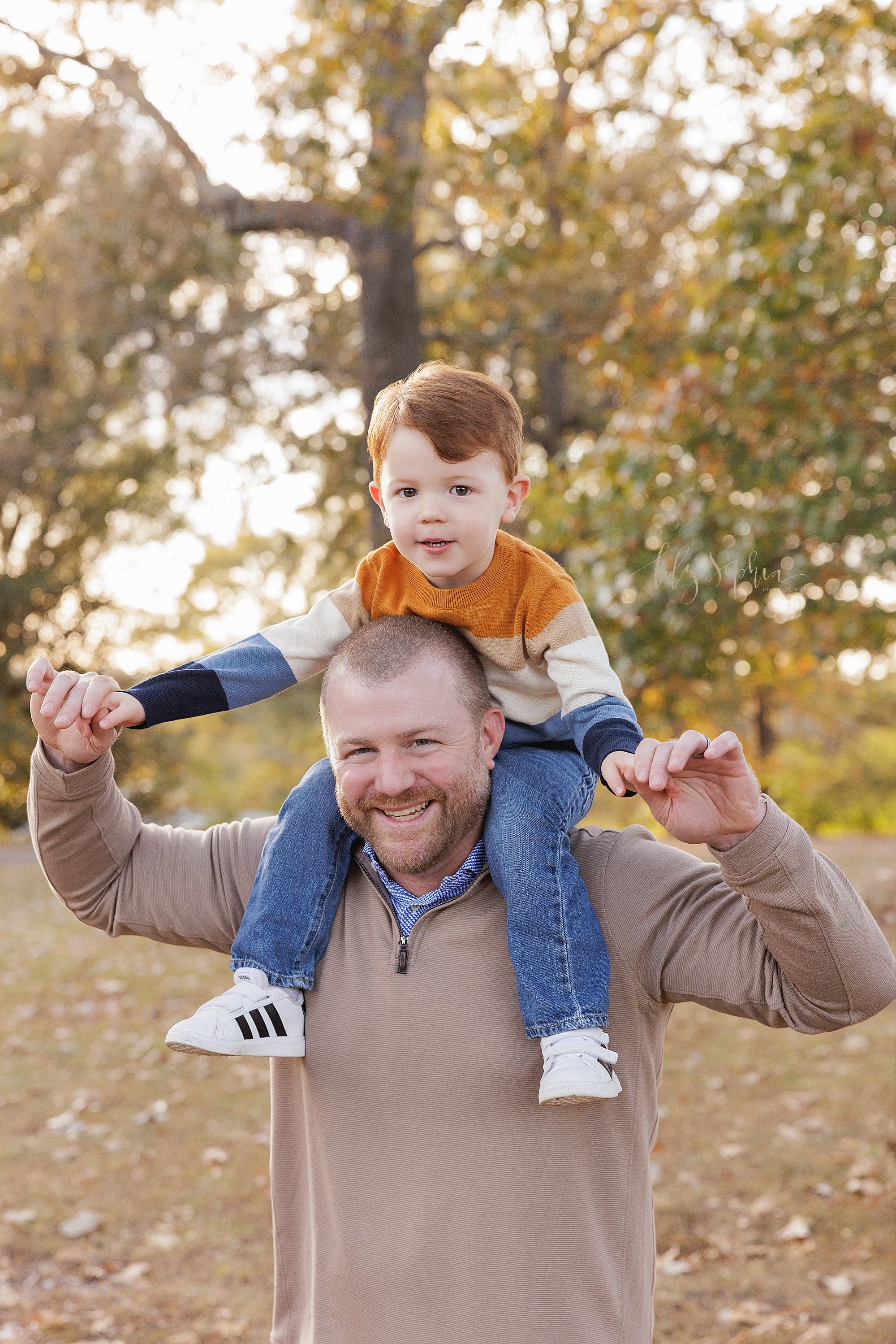  Family photo of a father carrying his young son on his shoulders as he holds his hands taken at sunset in a park near Atlanta, Georgia during autumn. 