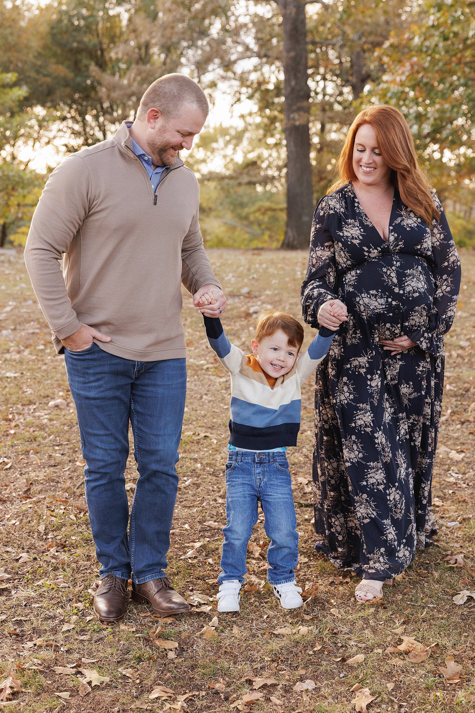  Family maternity portrait of a mother and father holding the hands of their toddler son as he walks between them with mom holding the base of her belly with her left hand taken during autumn in an Atlanta park at sunset. 