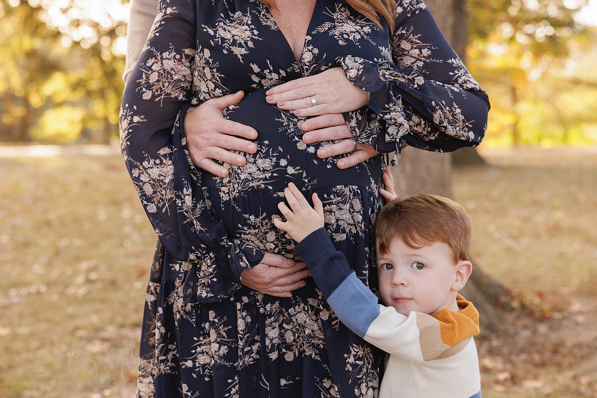 Maternity photo session with an expectant mother standing with her back to her husband’s chest as he wraps his hands around her to place them on their child in utero while mom is framing her belly with her hands and their toddler son stands on mom’s