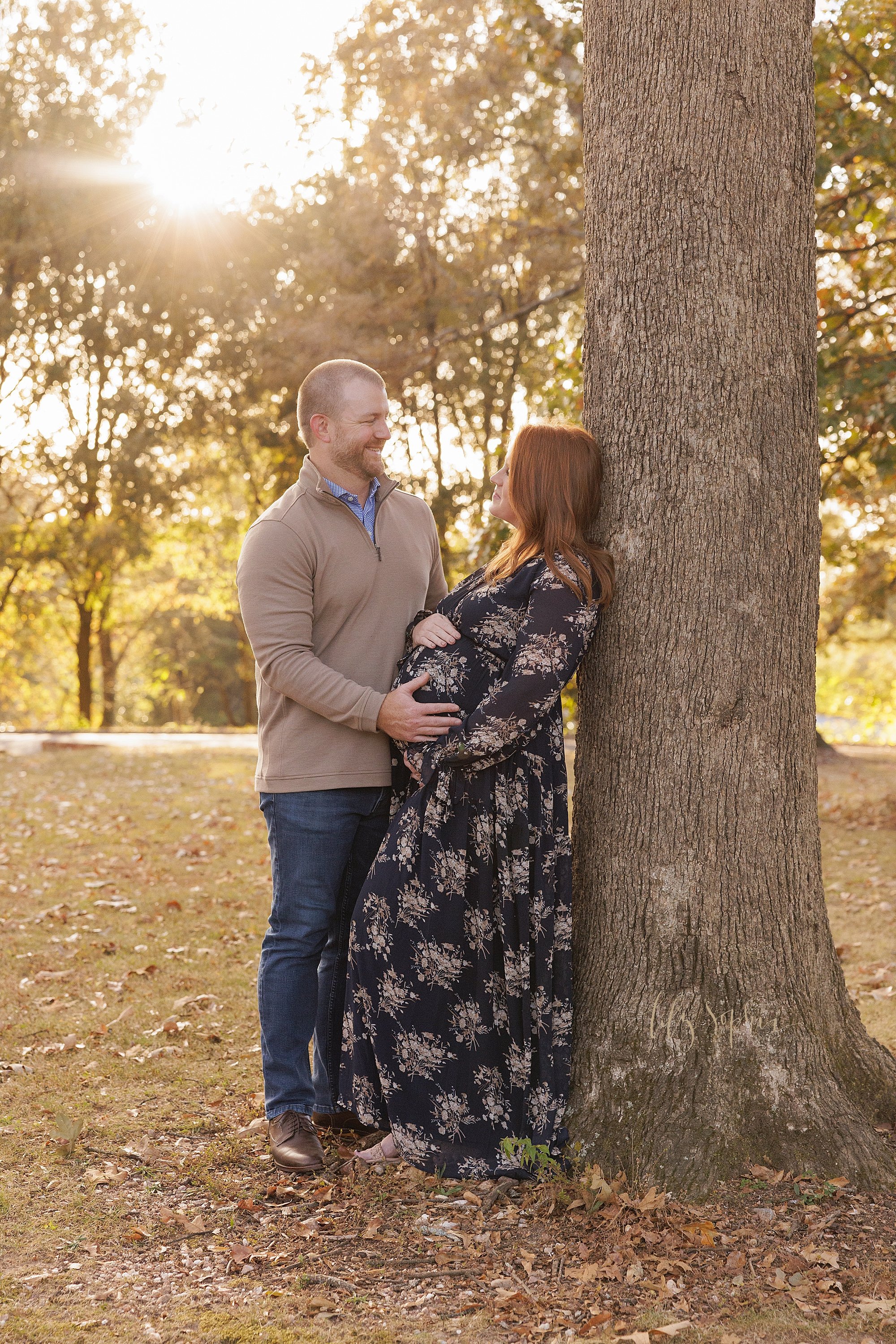  Maternity portrait of a pregnant mother standing with her back against a tree in an Atlanta park as her husband who is smiling at her faces her and places his hands on their child in utero taken at sunset during autumn. 