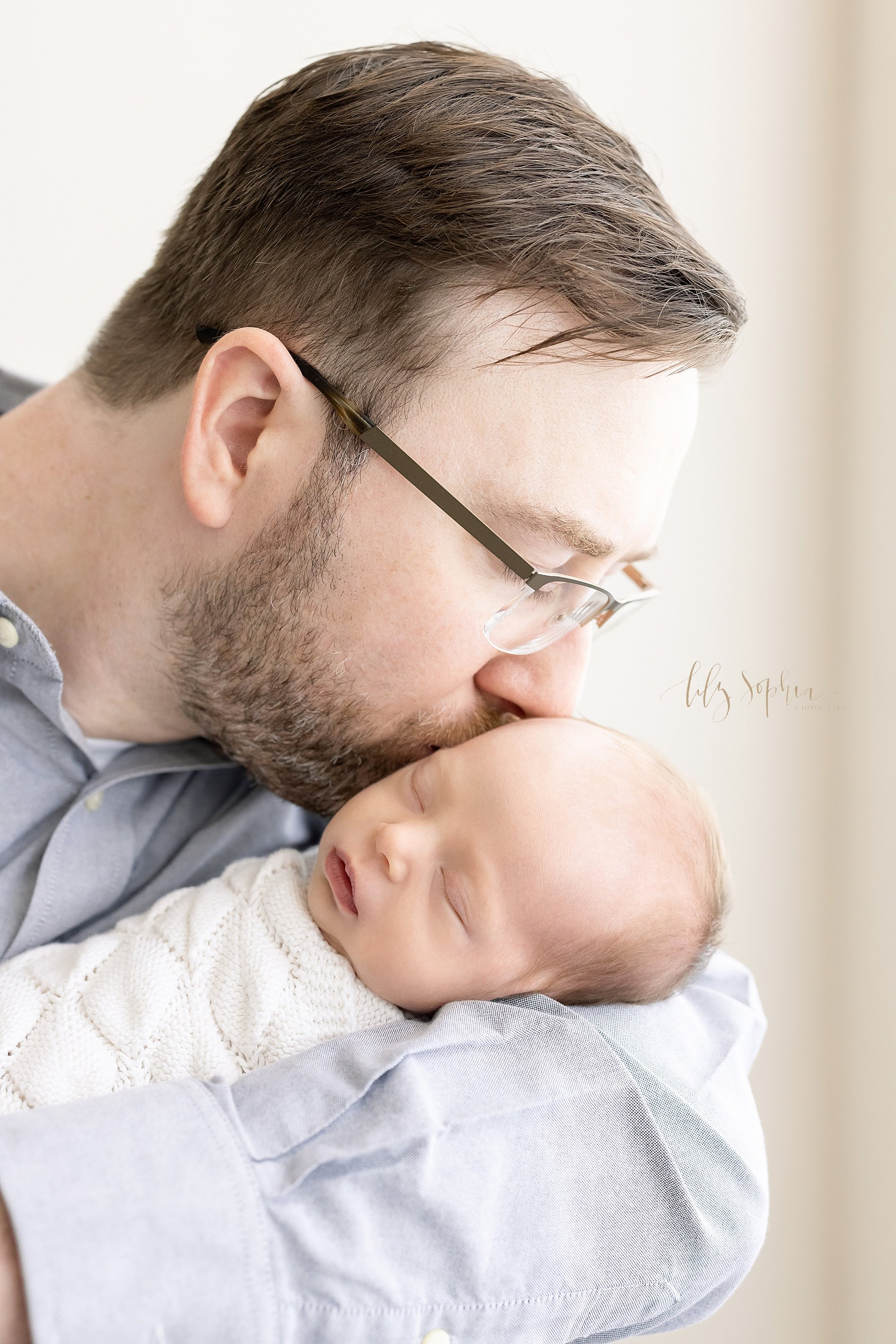  Newborn photo session with dad holding his newborn baby girl in his arms as he kisses the side of her head as she peacefully sleeps taken in a natural light studio near Brookhaven in Atlanta. 