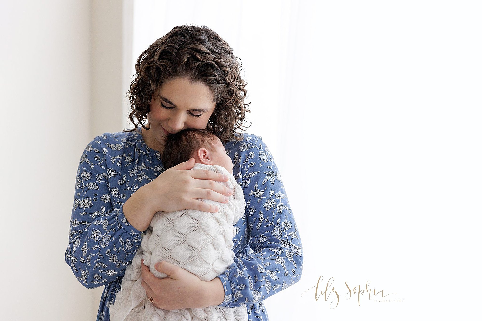  A mother cuddles her newborn baby girl against her chest treasuring these moments as she stands next to a window streaming natural light into a photography studio near Buckhead in Atlanta for a newborn photography session. 