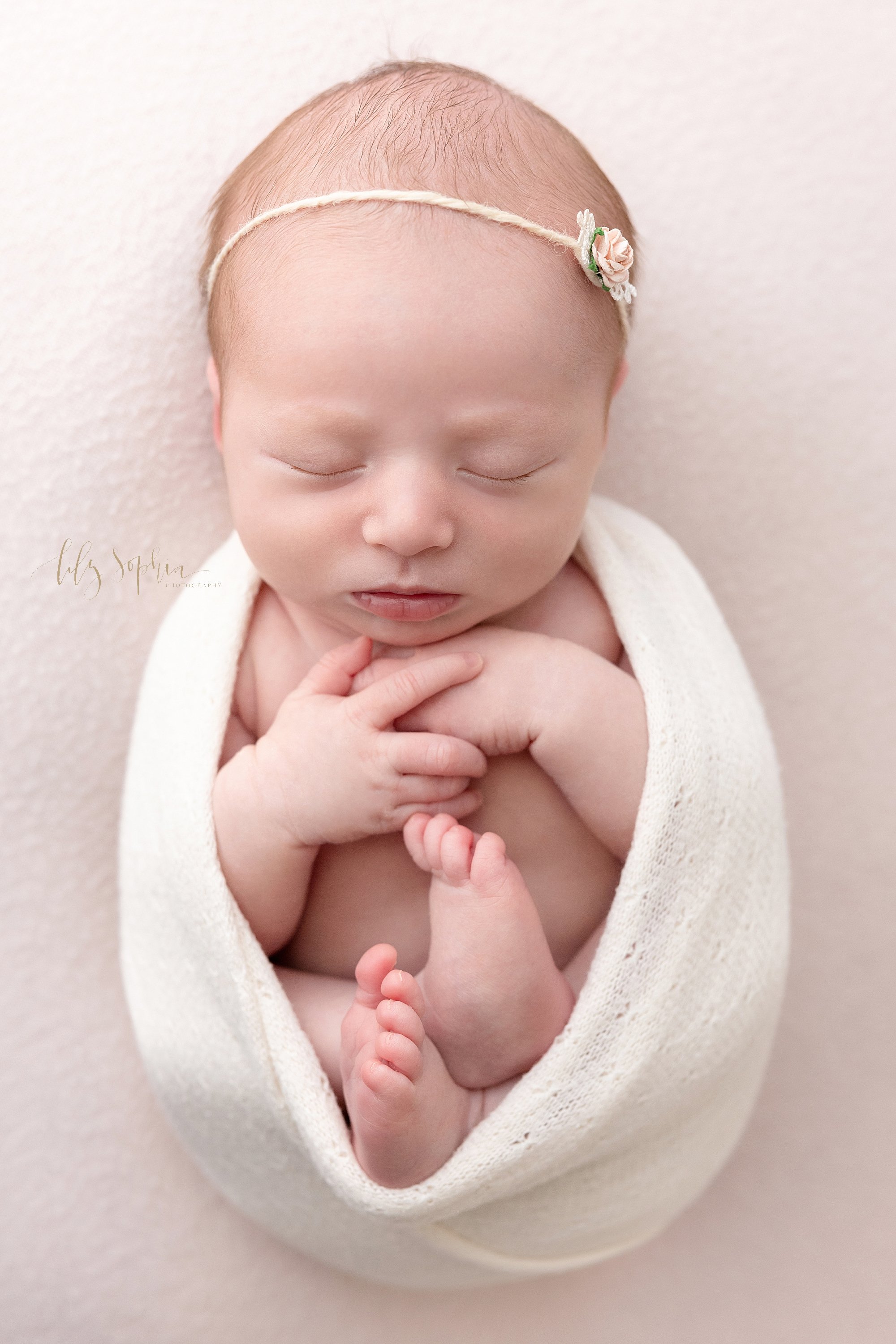  Newborn photo session with a newborn baby girl wearing a delicate rose headband in her wispy hair as she sleeps tucked into a stretchy swaddle taken in natural light in a photography studio near Buckhead in Atlanta. 