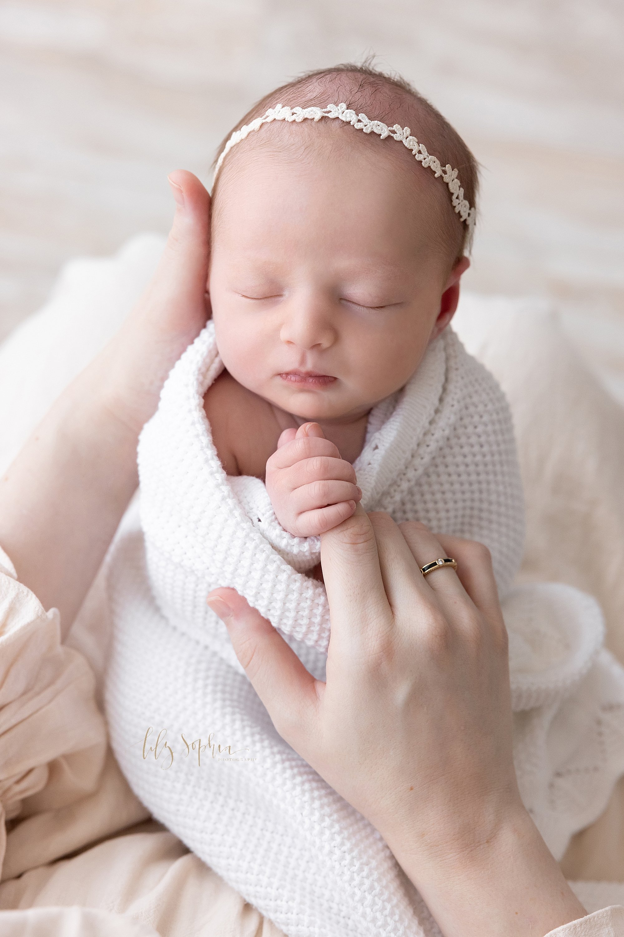  Newborn portrait of a newborn baby girl as she is held in her mother’s left hand and holds the pointer finger of her mother’s right hand as she peacefully sleeps taken near Kirkwood in Atlanta in a natural light photography studio. 