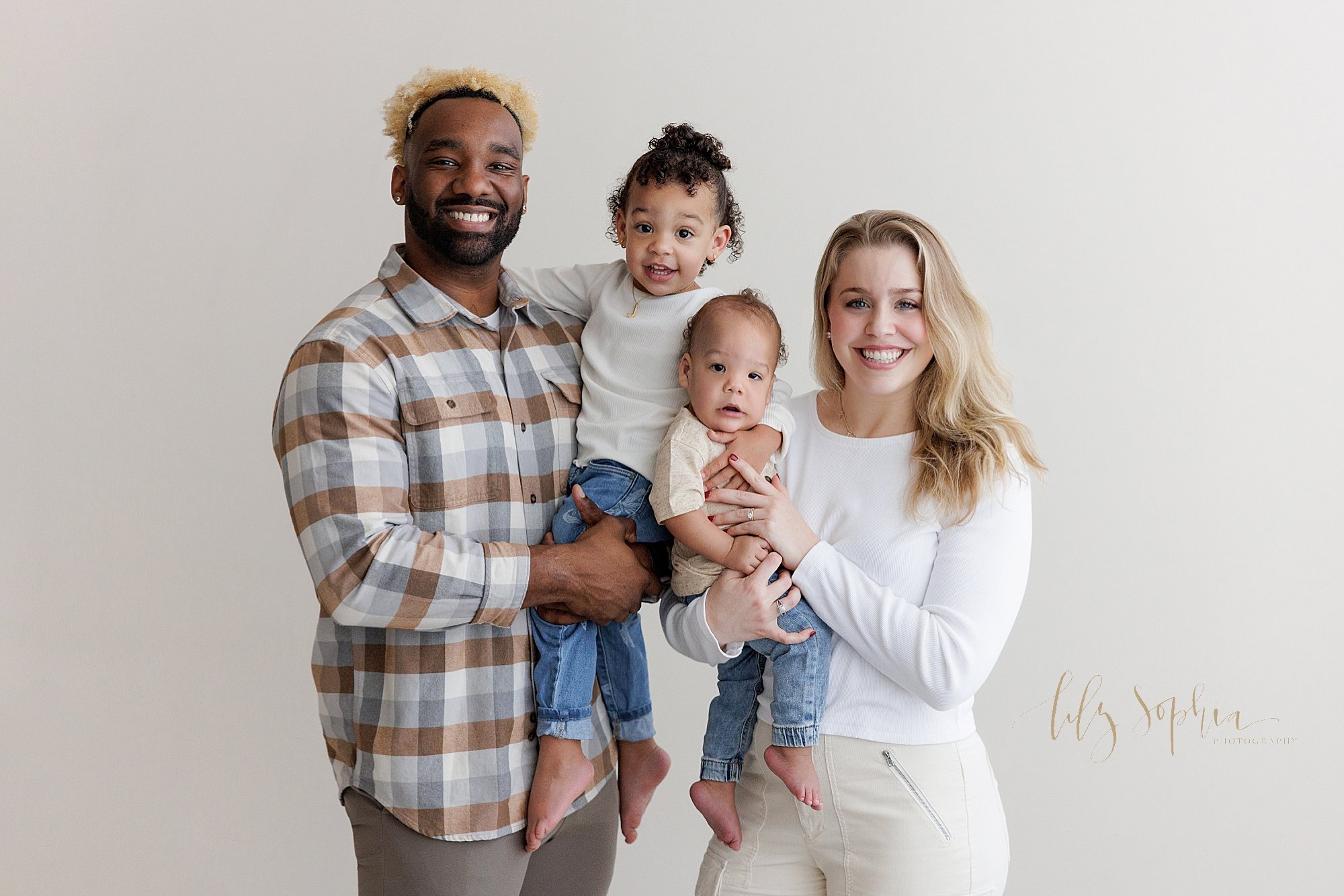  Family first birthday portrait of a father holding his toddler daughter in his arms as mom stands next to him holding their one year old son in her arms and his sister reaches to hold her brother taken near Poncey Highlands in Atlanta in a photograp