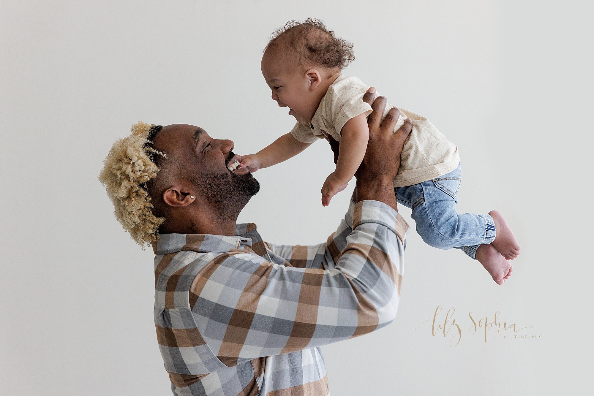  First birthday photo session with an African-American father holding his one year old son above his head as he laughs and reaches to touch his father’s face taken next to a window streaming natural light in a photography studio near Smyrna in Atlant