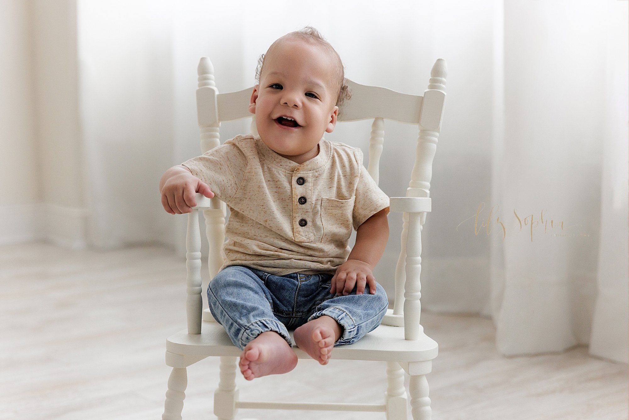  First birthday portrait of a one year old African-American boy as he rocks in a rocking chair in front of a window streaming natural light in a photography studio near Cumming in Atlanta, Georgia. 