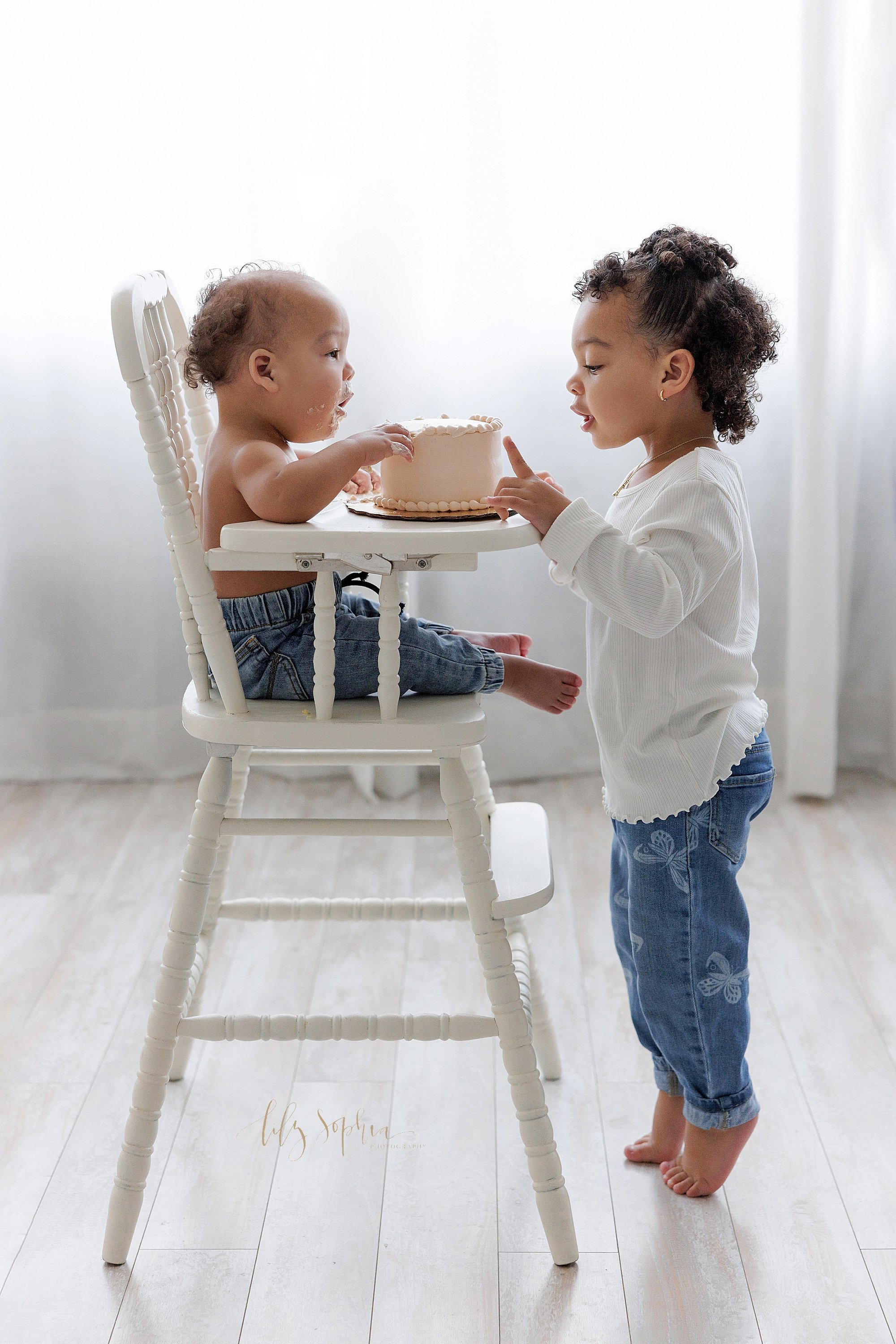  Sibling first birthday smash cake photo of a one year old boy sitting in an antique high chair with his smash cake on the tray as his sister stands on tiptoe in front of the high chair to give him instructions taken in a photography studio in front 
