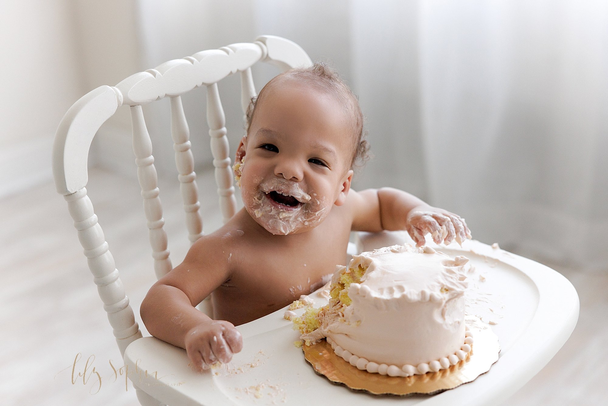  Smash cake photo of an African-American one year old boy as he sits in an antique wooden high chair after digging into his smash cake taken next to a window streaming natural light in Lily Sophia Photography Studio in Ponce City Market in Atlanta. 