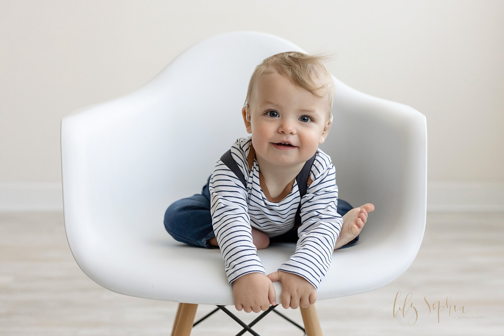  First birthday photo session of an active one year old baby boy as he sits in a white molded chair and reaches to hold on to the seat taken in natural light in a photo studio near Poncey Highlands in Atlanta. 