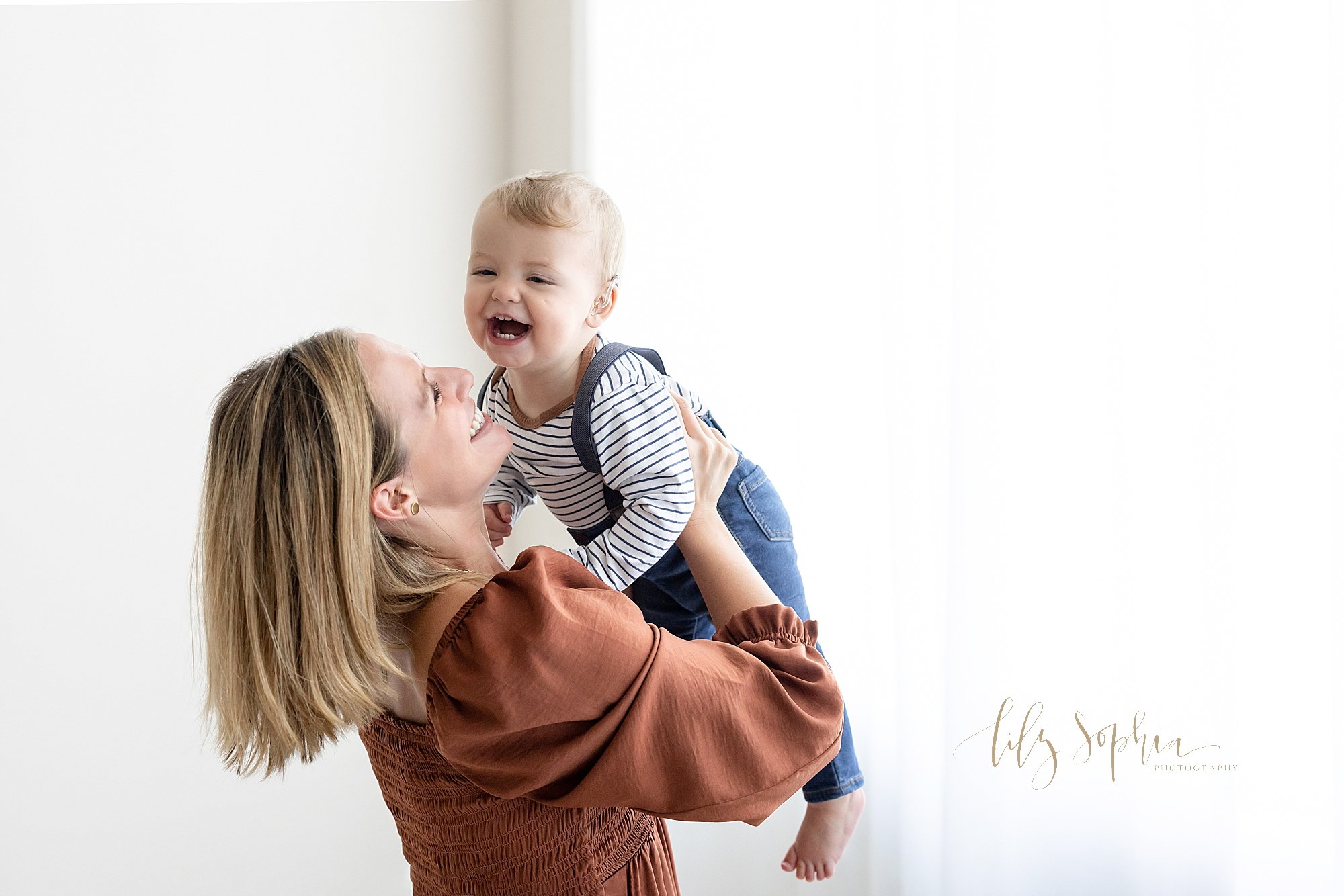  Mother and son share a fun moment as mom lifts her son above her and he giggles during a first birthday family photo session taken in front of a window streaming natural light in a studio near Sandy Springs in Atlanta. 