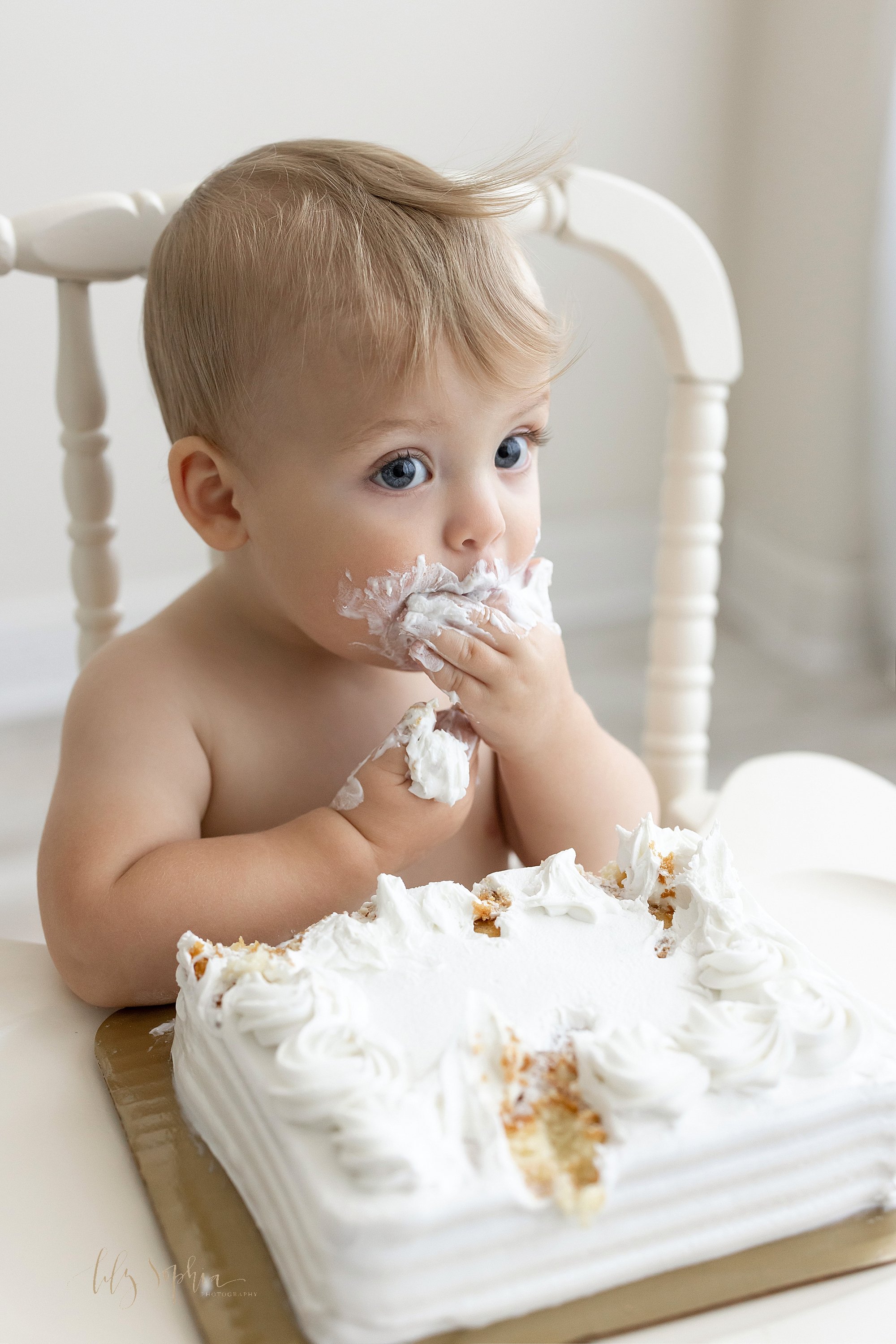  Smash cake one year old birthday photo of a baby boy as he sits in an antique high chair shoving icing in his mouth taken near Old Fourth Ward in Atlanta in a studio that uses natural light. 