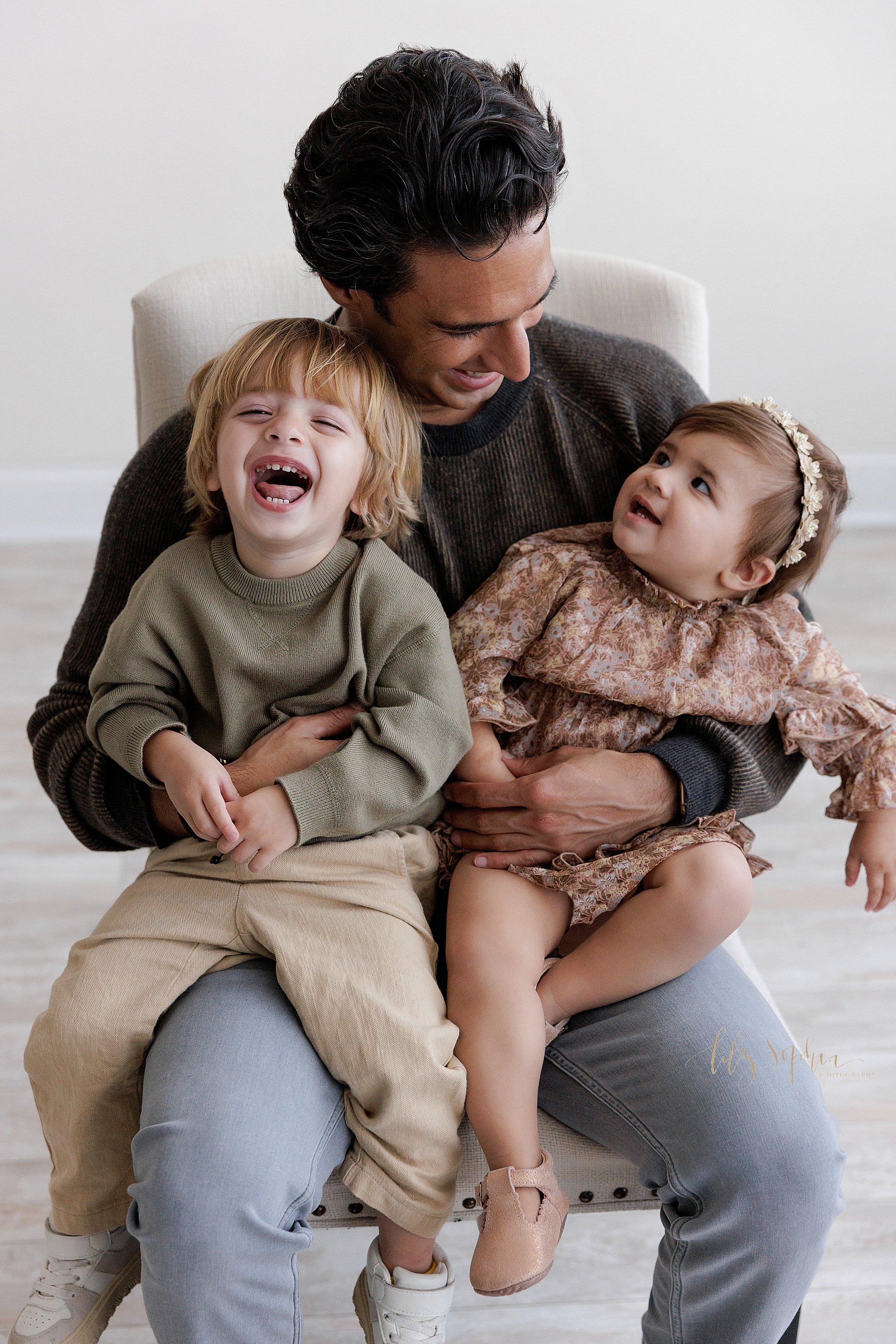  Family photo of a father holding his laughing toddler son on his right leg and his one year old daughter who is looking up to him on his left leg as he sits in a chair in a natural light studio near Virginia Highlands in Atlanta. 