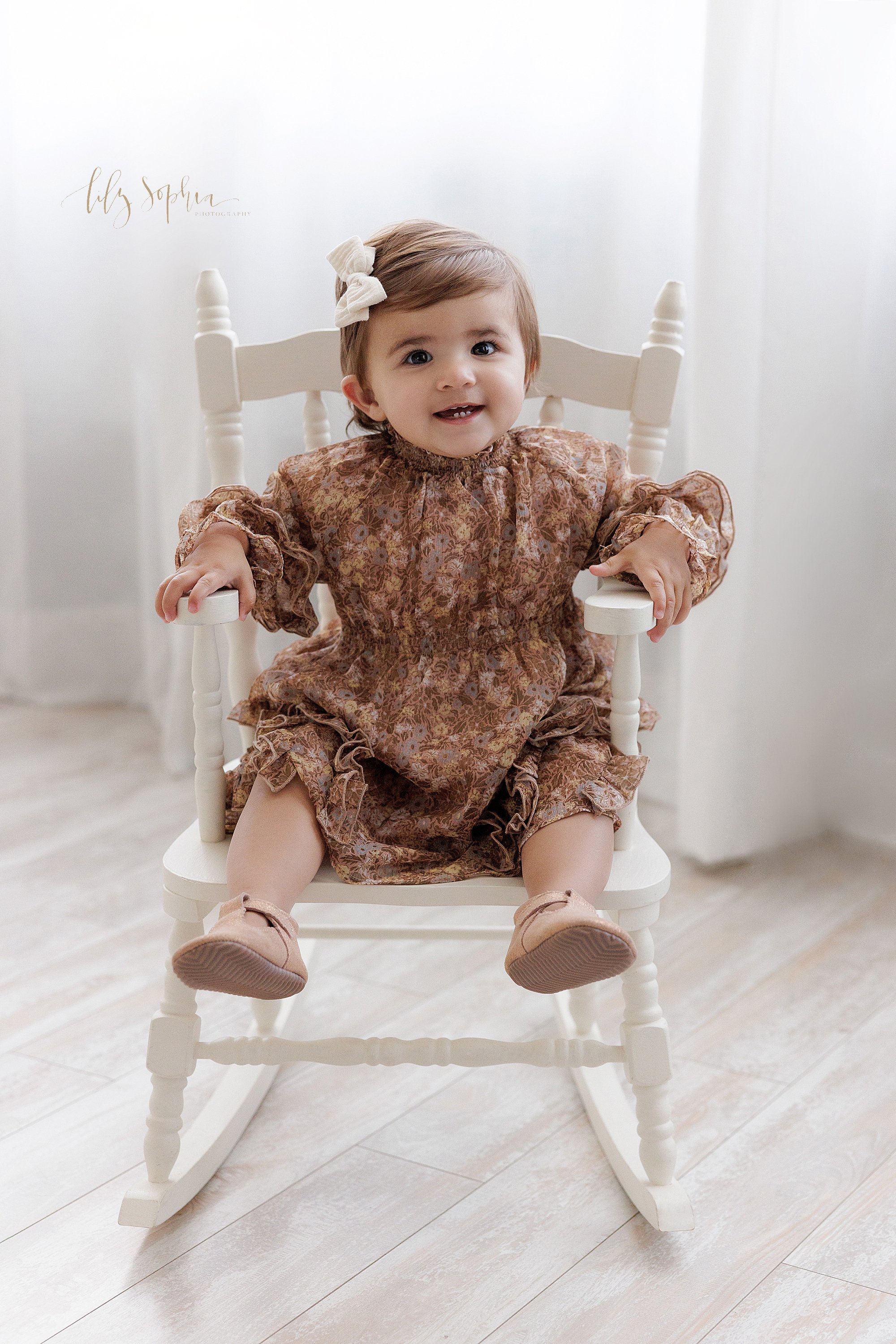  First birthday portrait of a one year old little girl as she smiles while sitting in a rocking chair in front of a window streaming natural light in a photography studio near Oakhurst in Atlanta. 