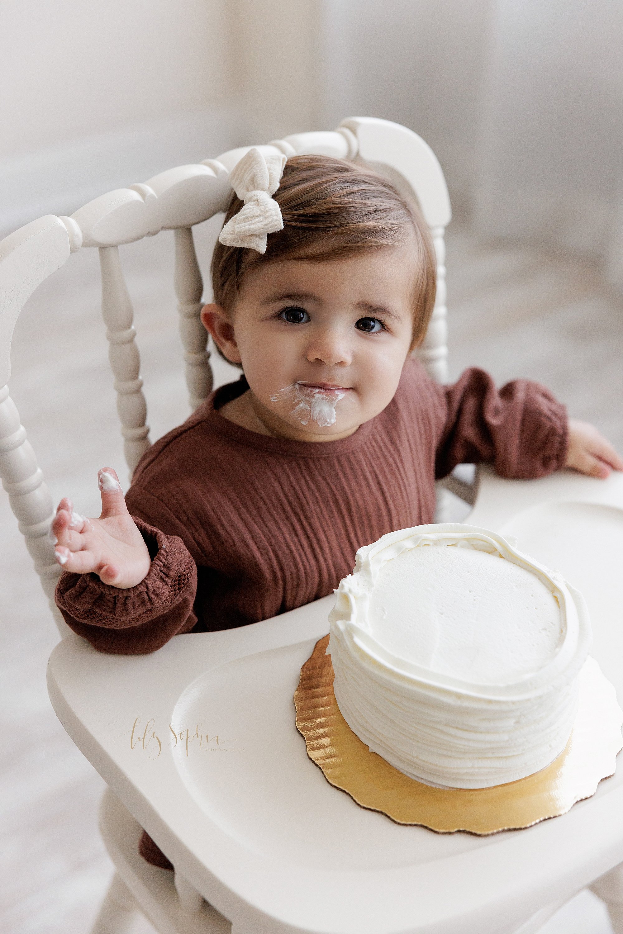  First birthday smash cake portrait of a one year old little girl as she sits in an antique high chair with icing on her right hand and her face taken near Old Fourth Ward in Atlanta in a natural light studio. 