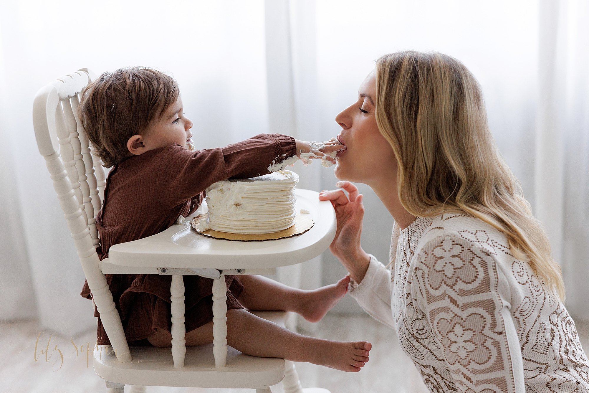  First birthday smash cake photo session with the one year old little girl sitting in an antique high chair as she feeds icing to her mother who is squatting in front of her taken in  front of a window streaming natural light in a photography studio 