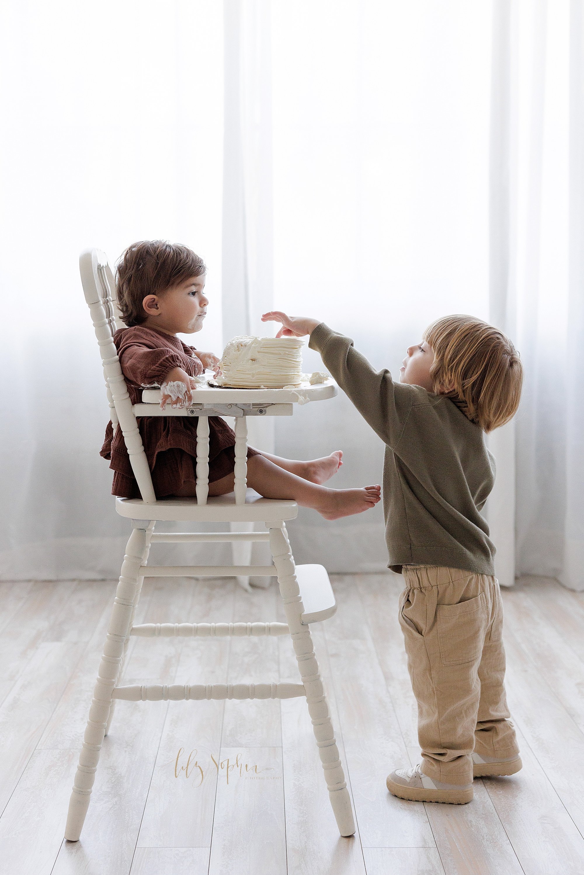 Sibling smash cake photo of a one year old girl sitting in an antique high chair with her smash cake on the tray watching her older brother reach to get a bite of icing taken in front of a window streaming natural light near Buckhead in Atlanta in a