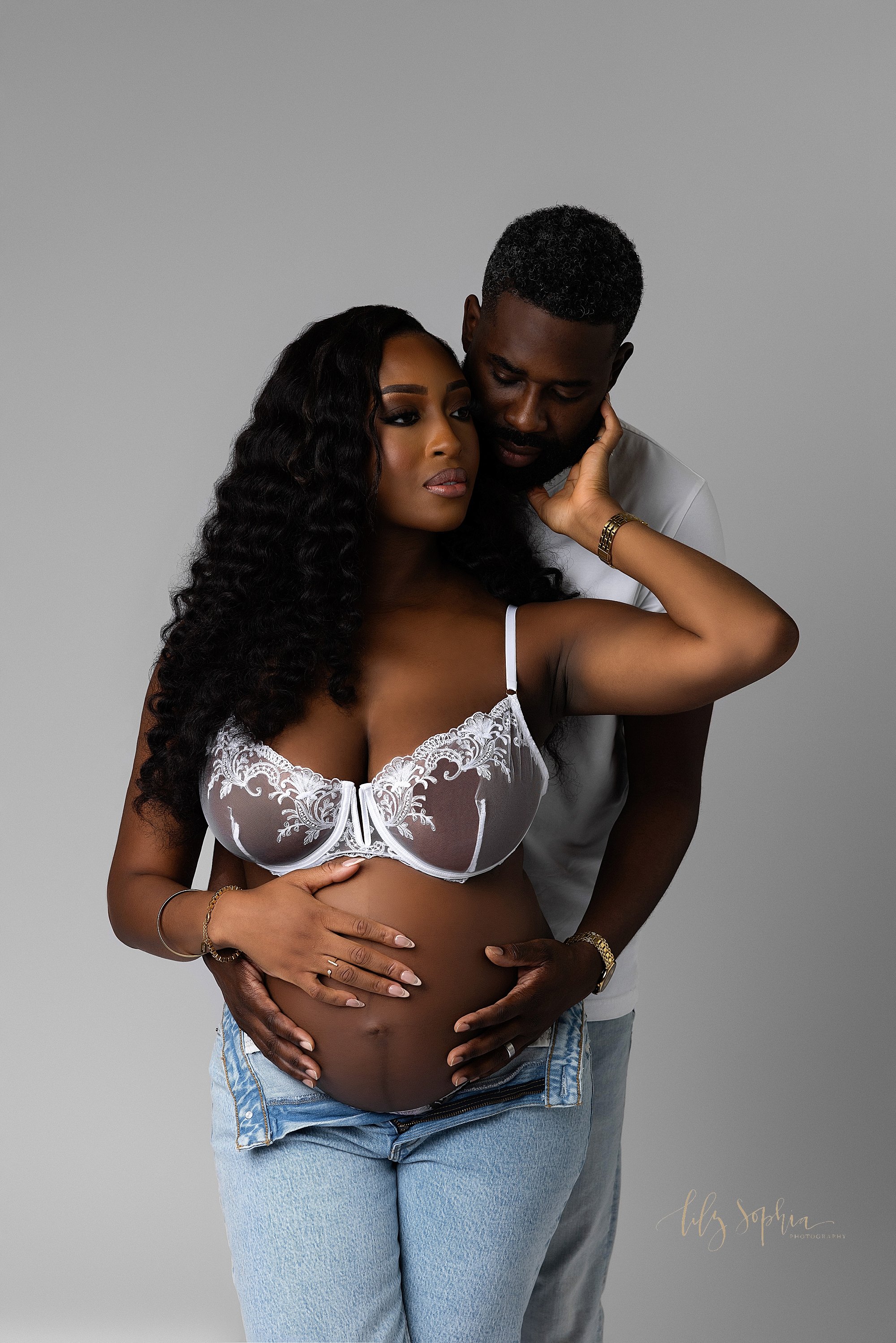  Modern maternity session with an African-American expectant mother wearing a white bra and unzipped blue jeans as her husband stands behind her and wraps his arms around her to place his hands on their child in utero as she reaches her left hand up 