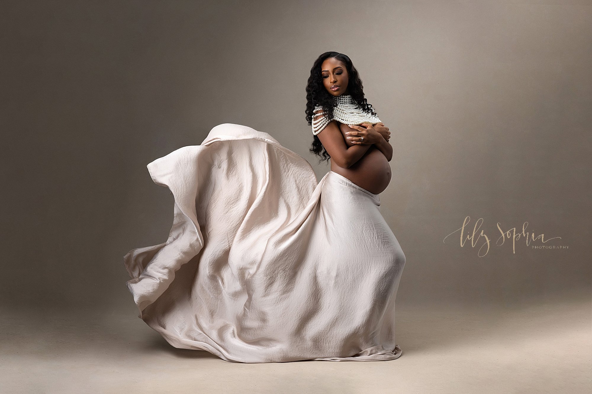  Modern maternity photo shoot with a pregnant African-American woman wearing a silky skirt and a beaded collar with the skirt billowing behind her taken near Decatur in Atlanta, Georgia in a photography studio. 
