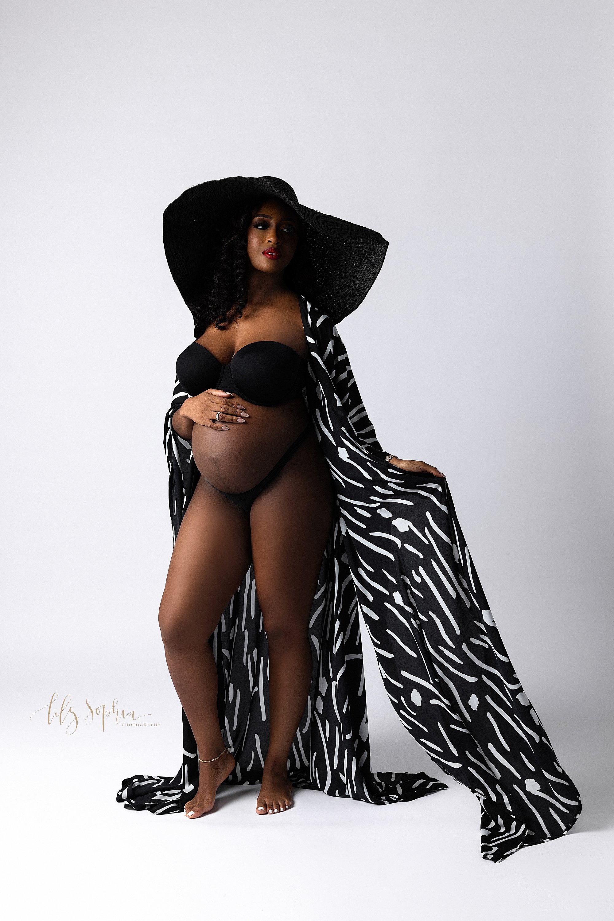  Modern maternity photo session with an African-American expectant mother wearing a wide-brimmed black hat, black underwear and a black and white silk robe as she places her right hand on her belly and uses her left hand to hold the robe open as she 