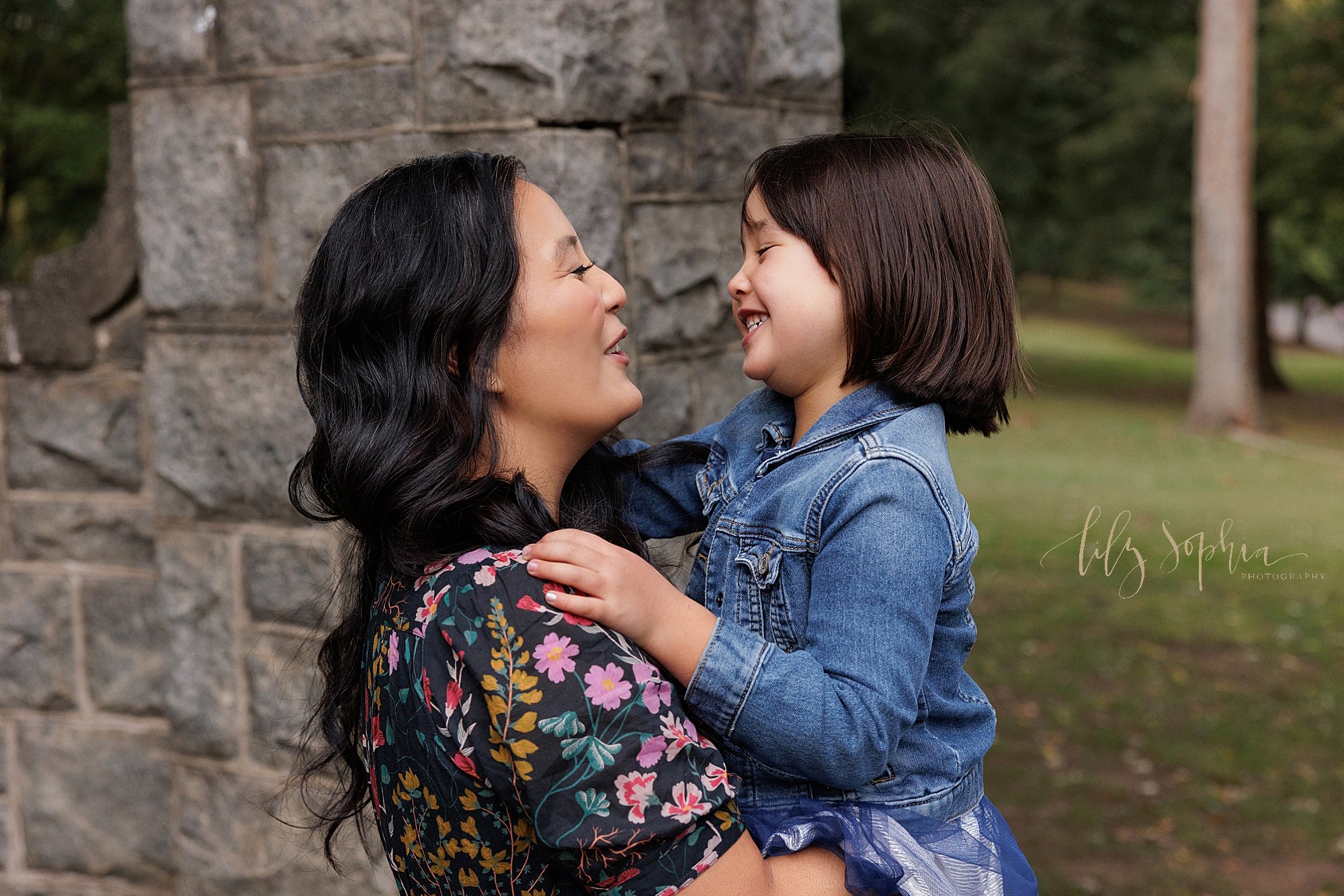  Family picture of a mother holding her young daughter in her arms as they talk to one another while mom stands next to a stone entranceway in a park at sunset during autumn in Atlanta, Geogia. 