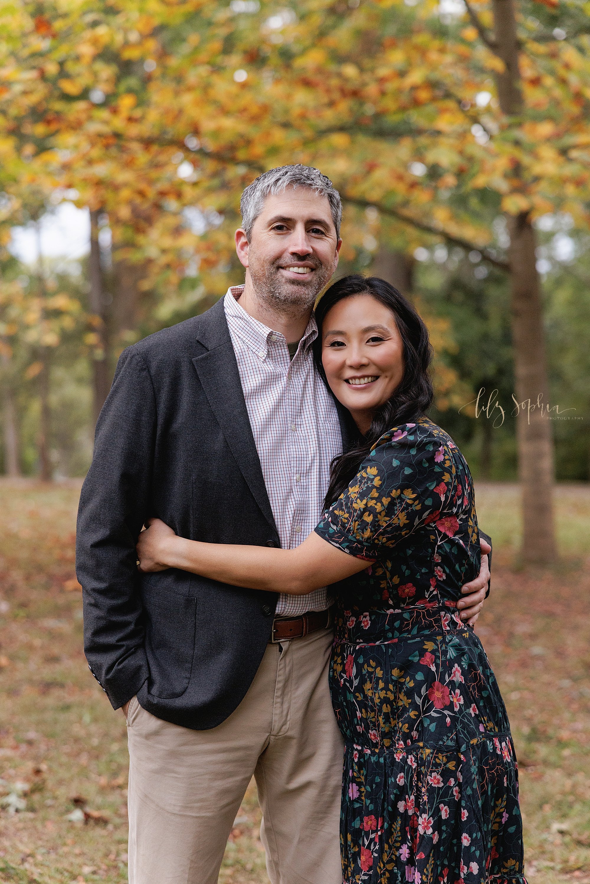  Family photo of a husband and wife with the wife hugging her husband as they stand in a park at sunset near Atlanta among the fall leaves. 